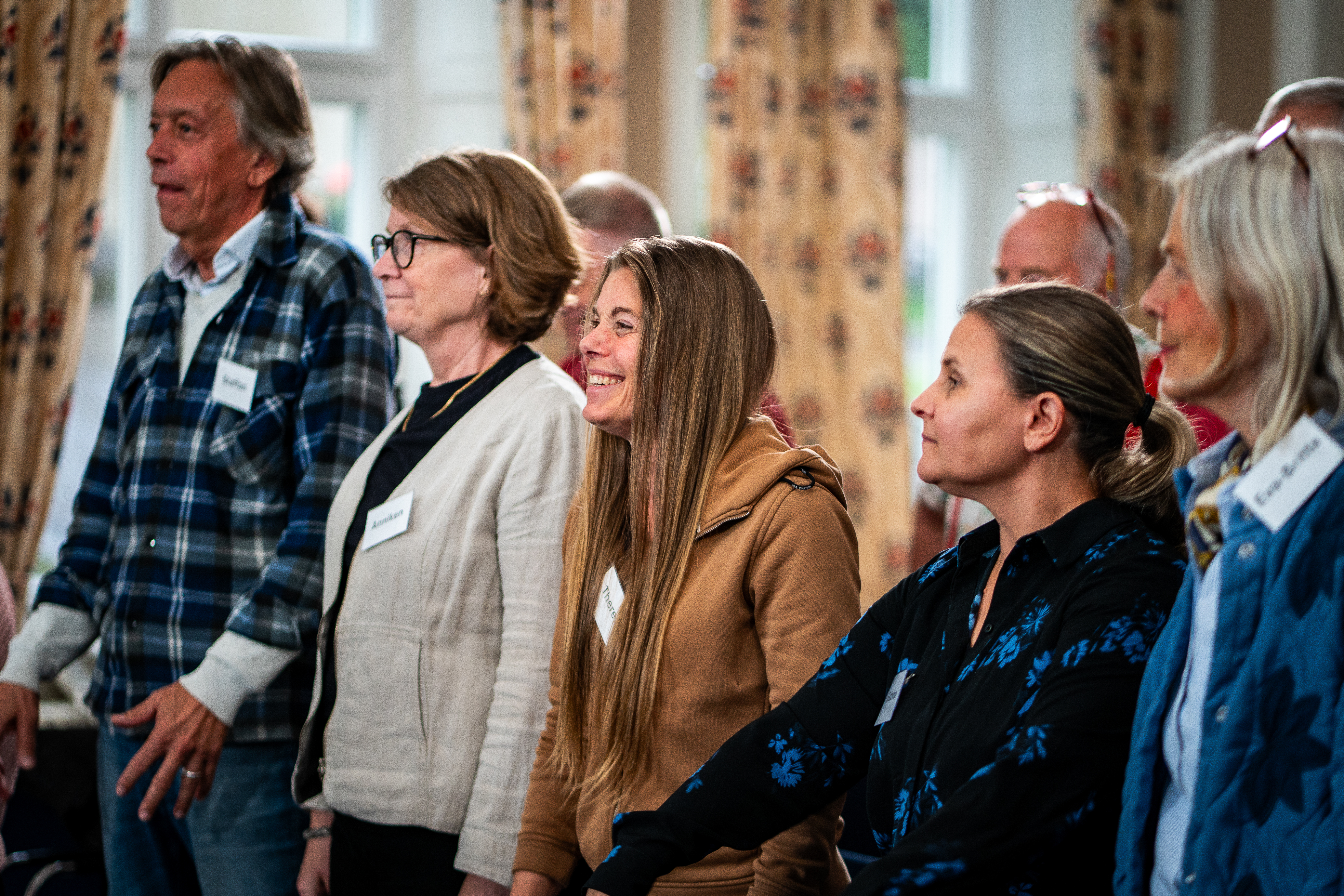 En grupp vuxna, som är deltagare i demenskören, står inomhus och sjunger. Några av dem tittar framåt och ler.