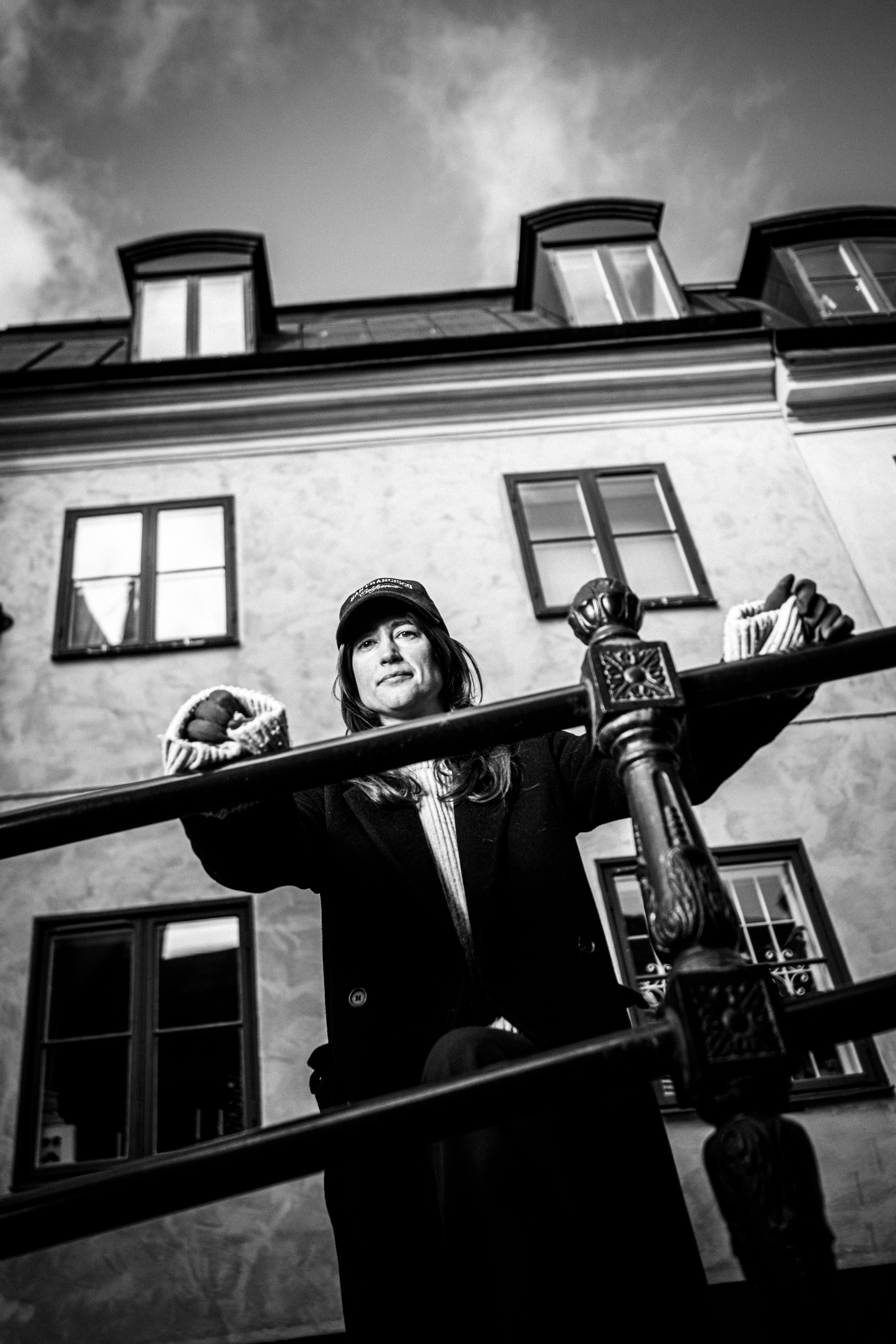 En person med rock och hatt står bakom ett räcke framför en byggnad med flera fönster.