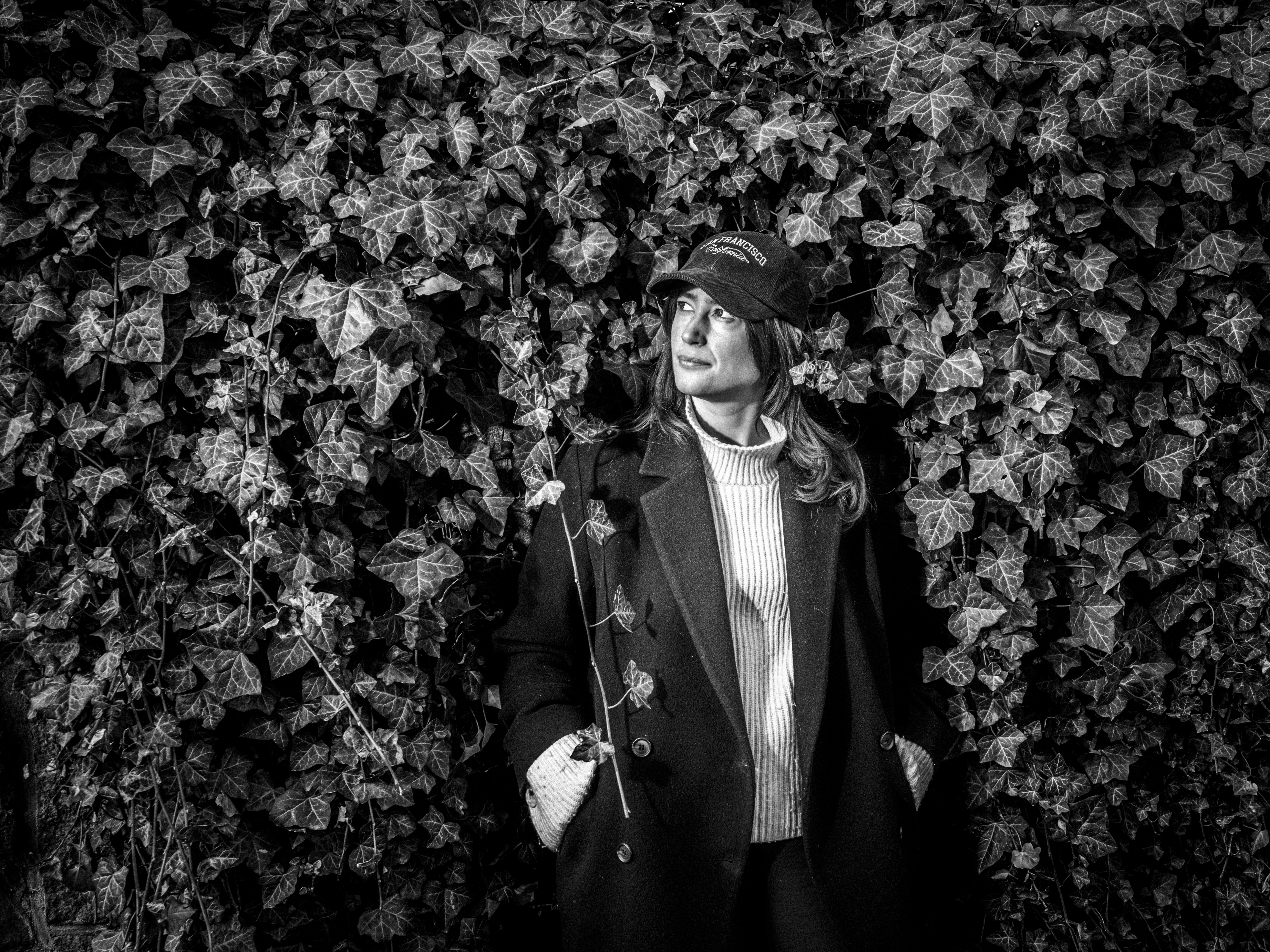 En person med rock och mössa står framför en vägg av murgröna och tittar åt sidan. Bilden är i svartvitt.