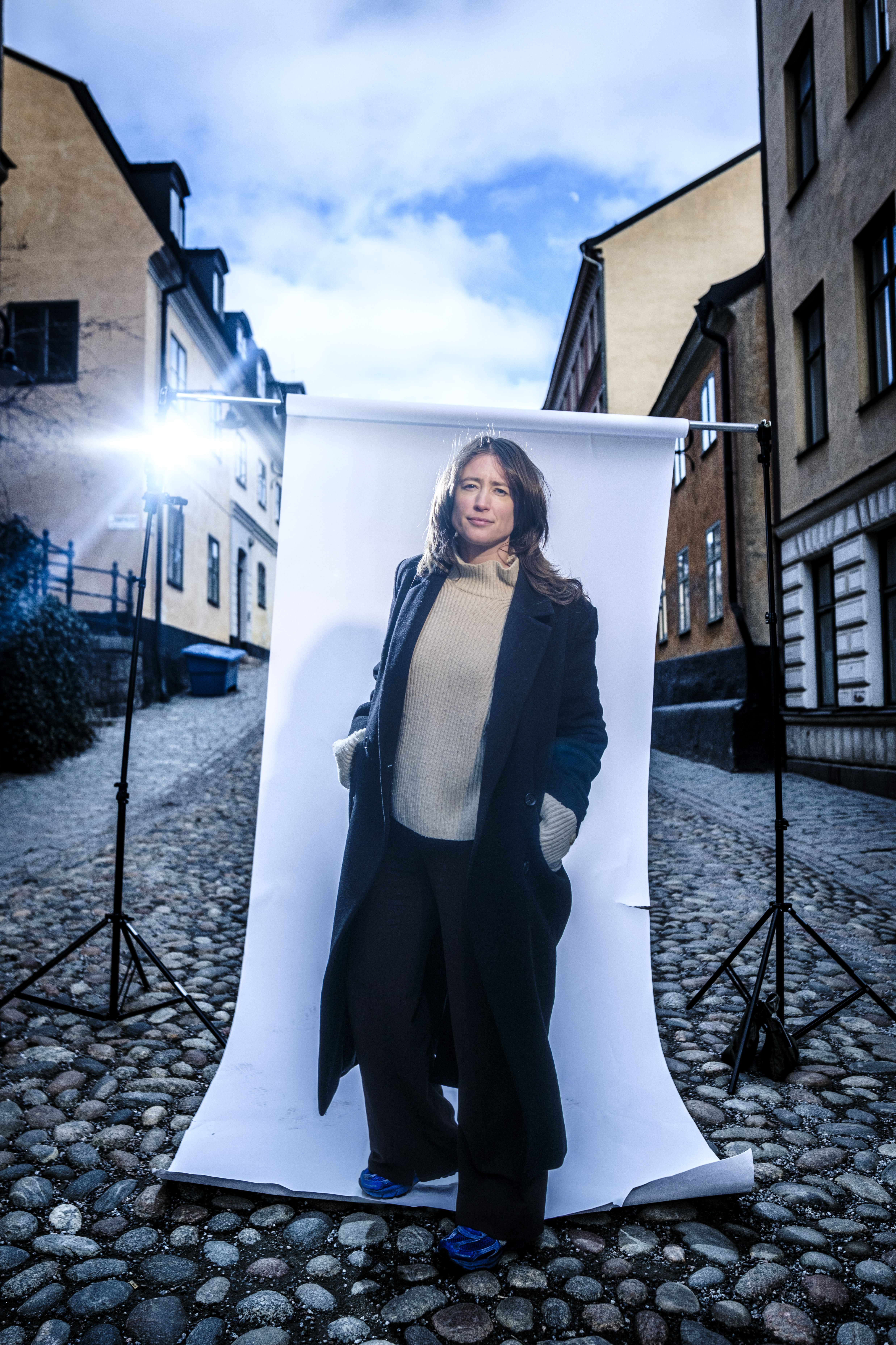 En person står på en gatstensbelagd gata framför en vit bakgrund, med belysningsutrustning och byggnader i bakgrunden.