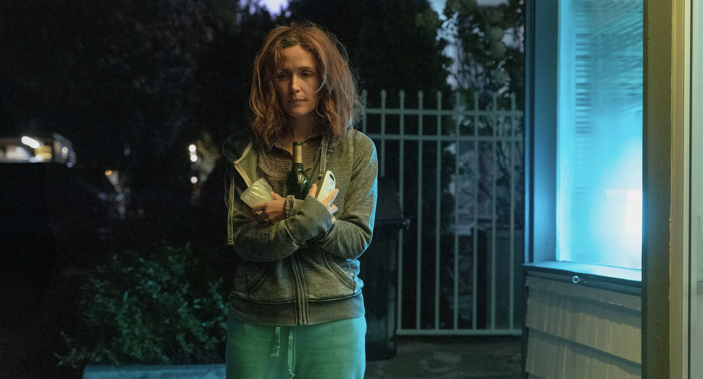 En kvinna med rufsigt hår i en grå huvtröja står utomhus på natten, håller flaskor och snacks nära ett upplyst fönster.
