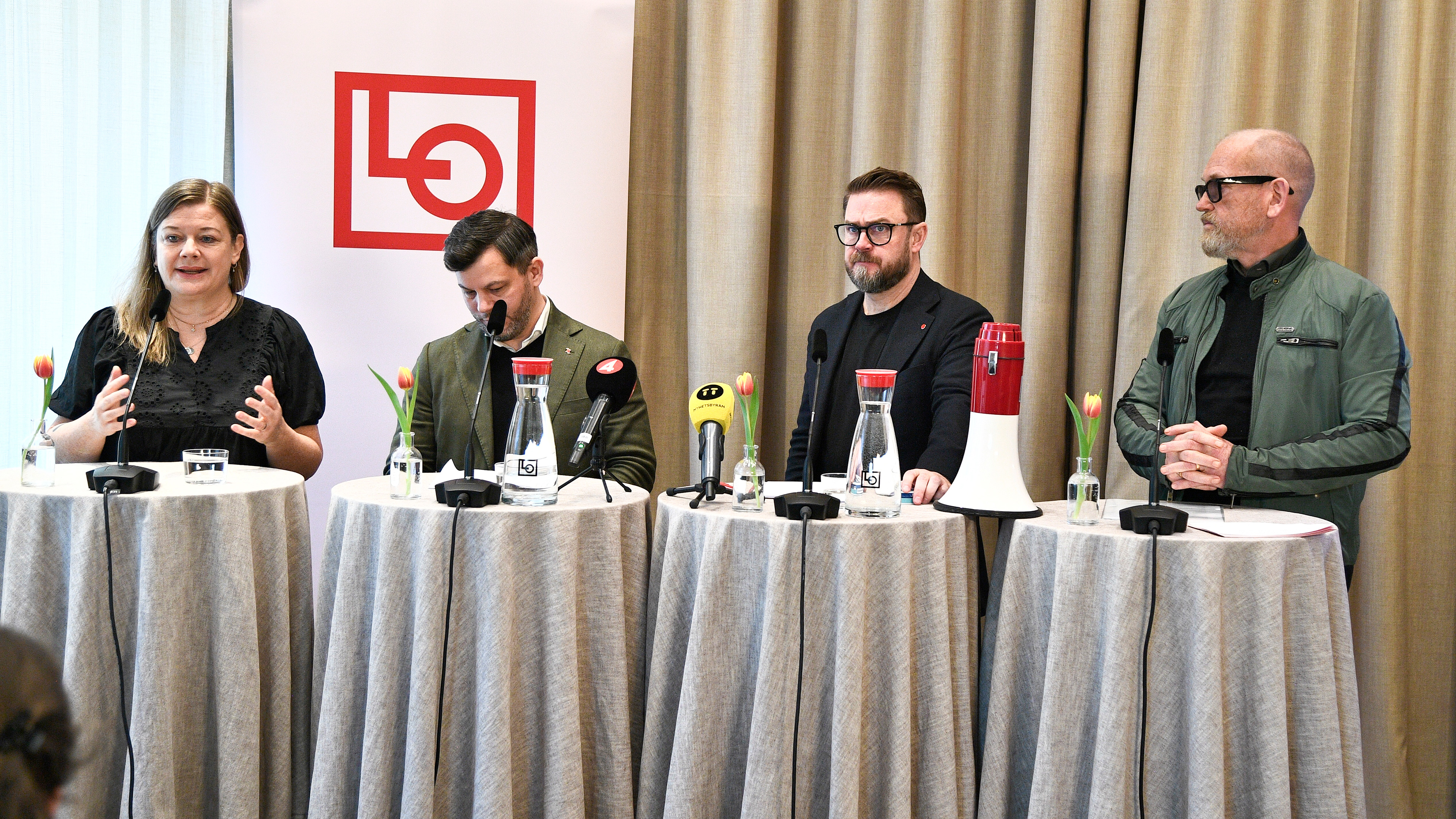 Fyra personer står bakom runda bord med mikrofoner och vattenflaskor vid en pressträff framför en LO-logga och beige gardiner.