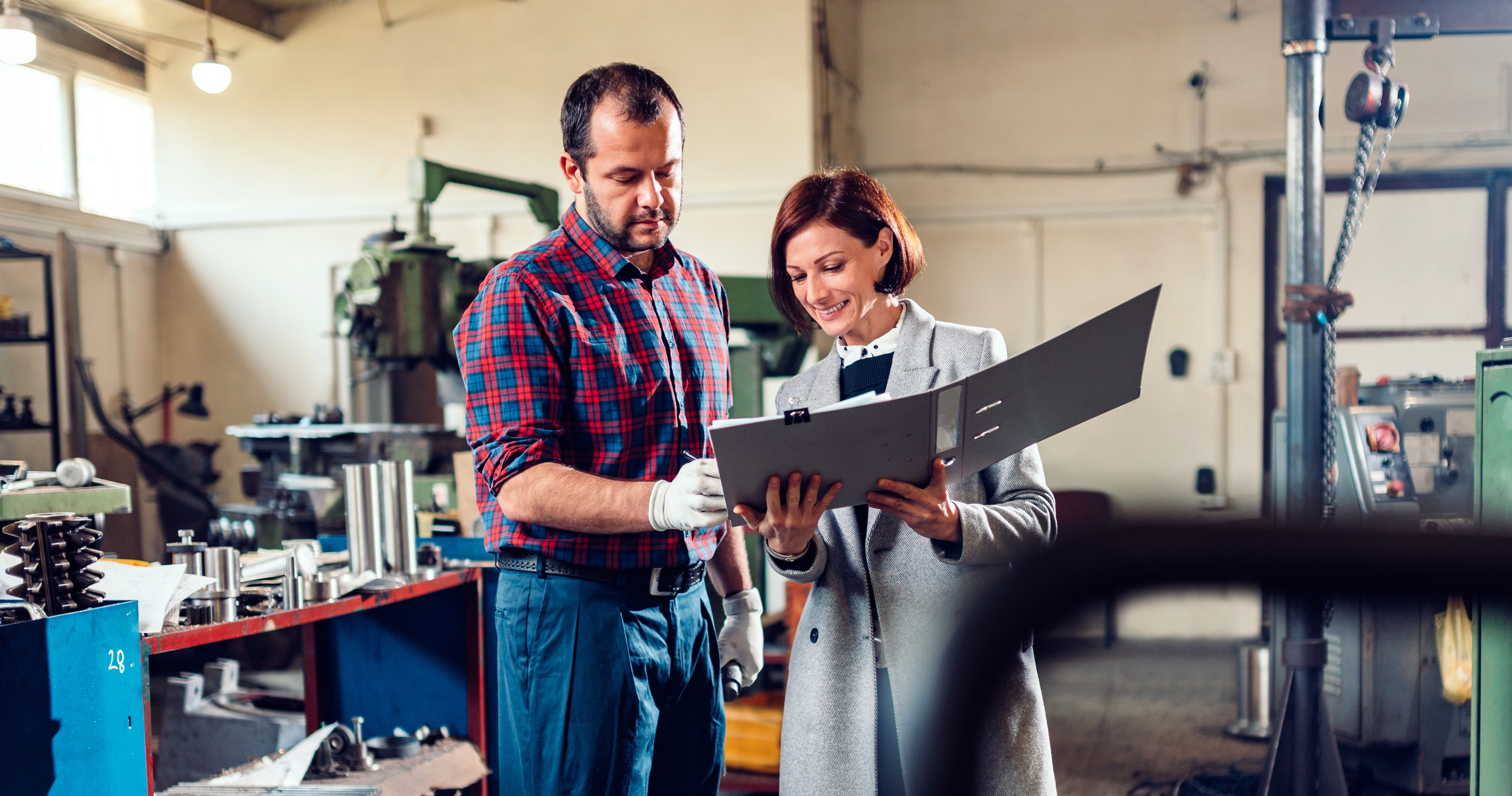 En man med arbetskläder och handskar och en kvinna med rock går igenom dokument tillsammans i en verkstad med maskiner och verktyg.