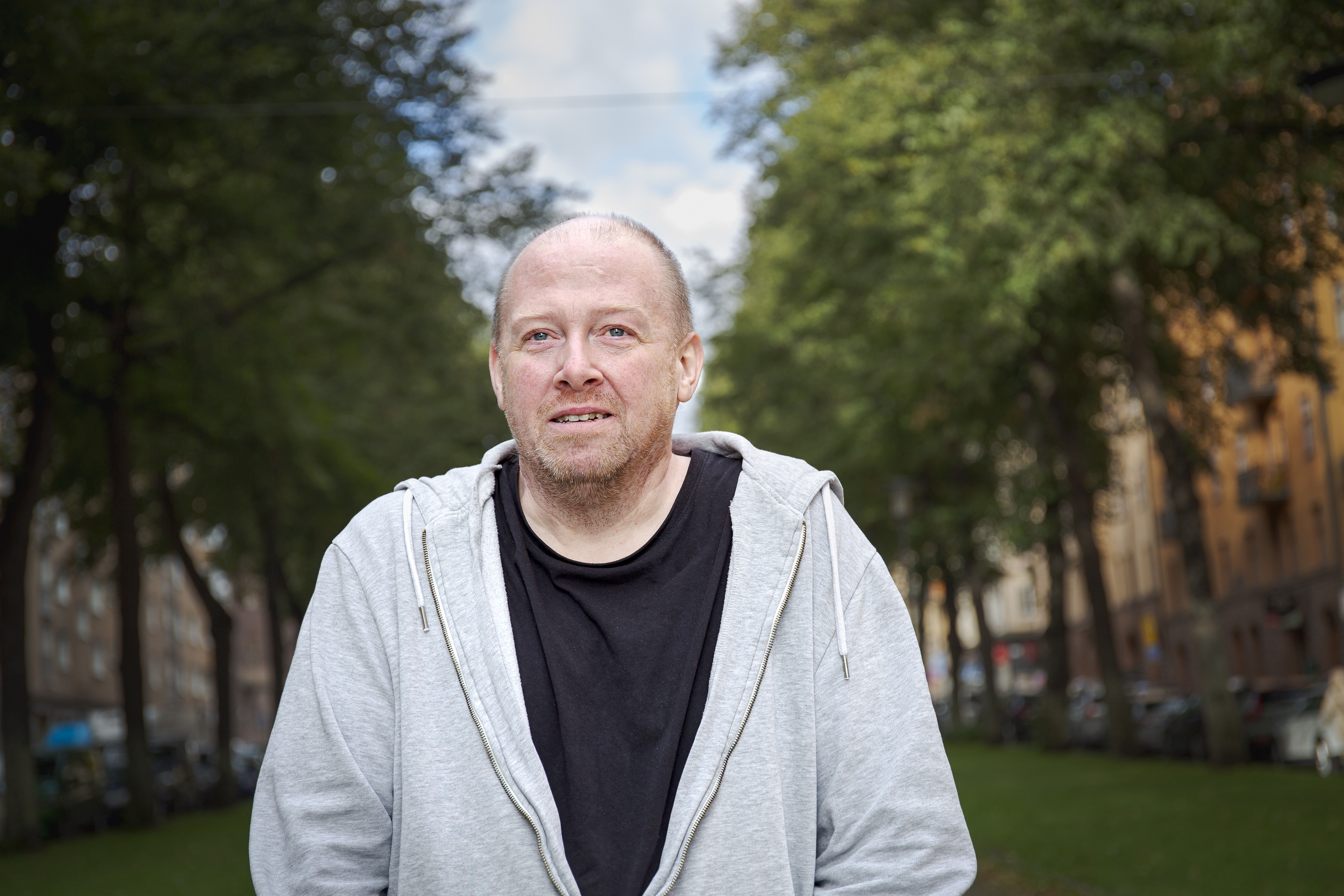 Daniel Pallin med grå huvtröja och svart tröja står utomhus på en gata med träd, byggnader och parkerade bilar i bakgrunden.