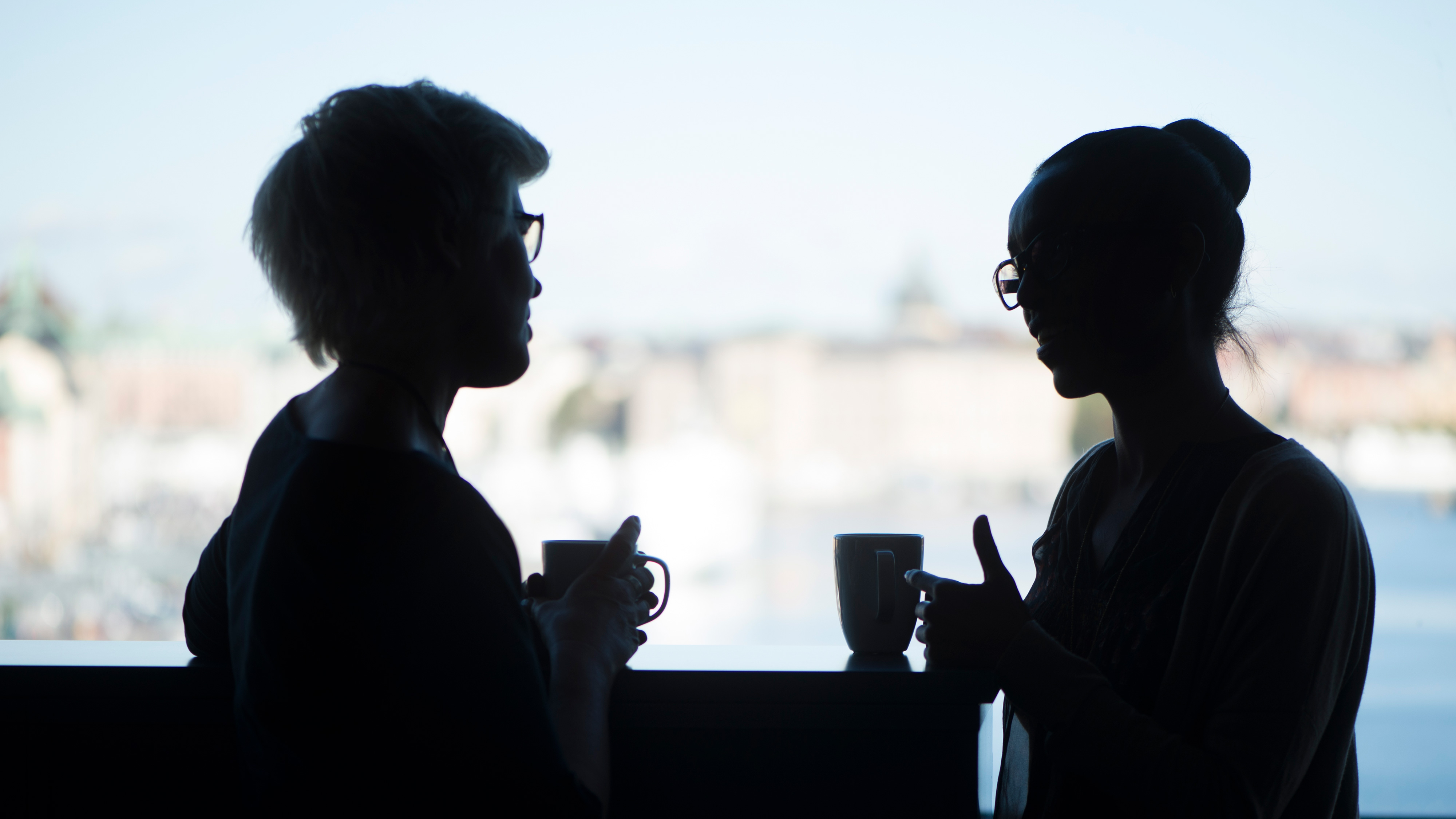 Två personer i silhuett håller varsin mugg och samtalar vid ett fönster med en suddig stadsmiljö i bakgrunden.