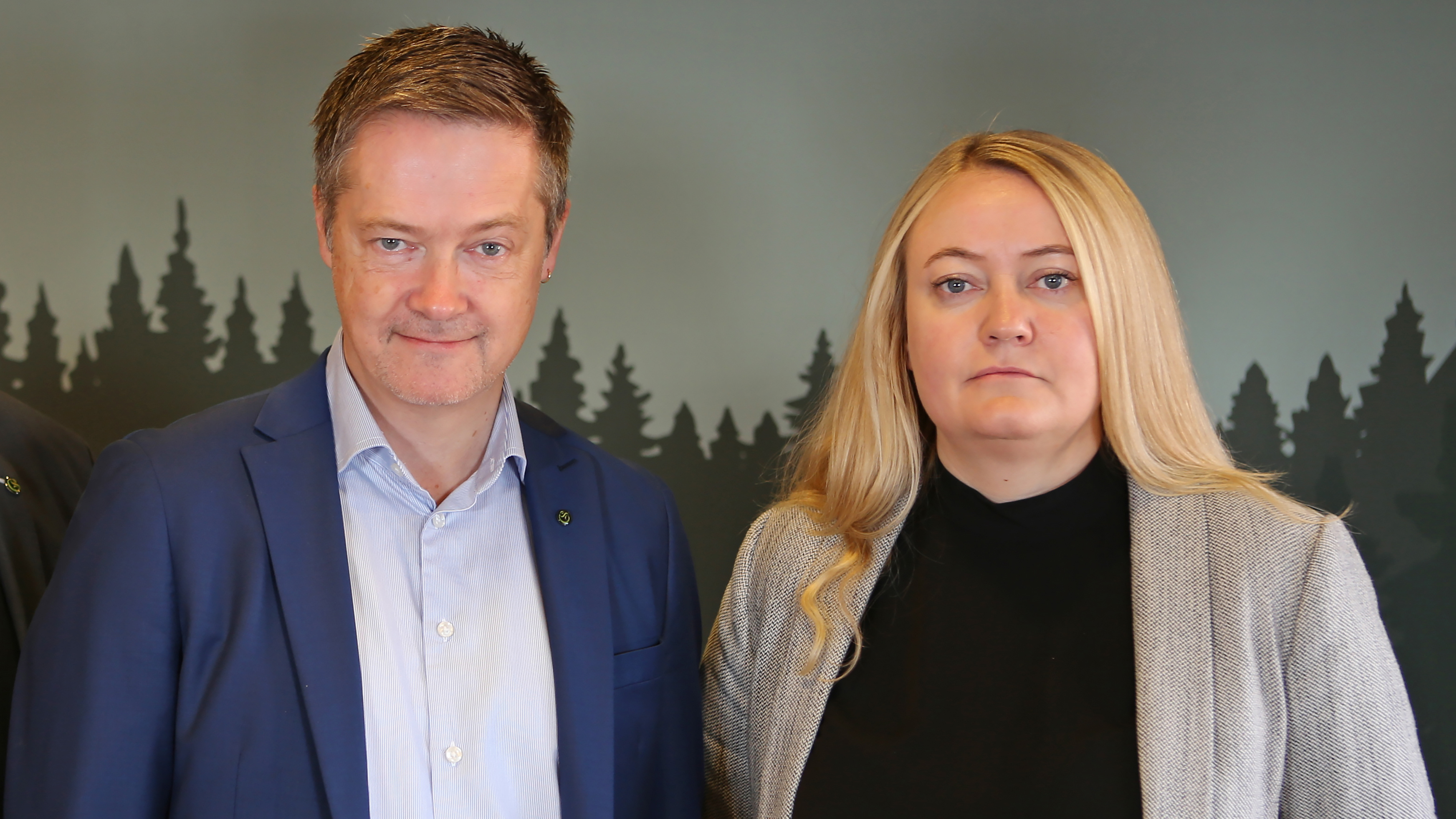 Två personer, en man i blå kostym och en kvinna i grå kavaj, står bredvid varandra framför en bakgrund med trädmönster.