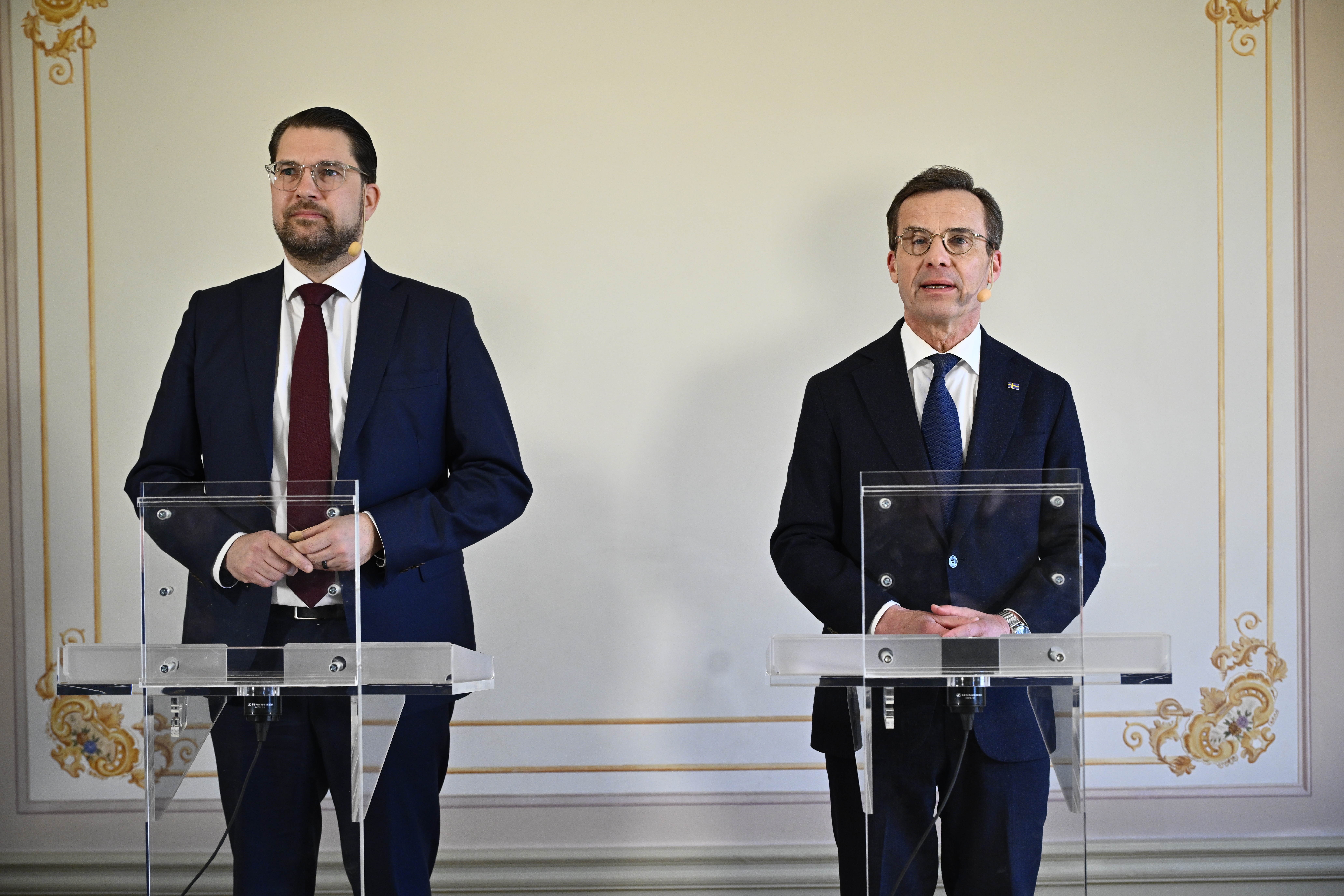 Två män i kostym står framför en vägg med gamla mönster.