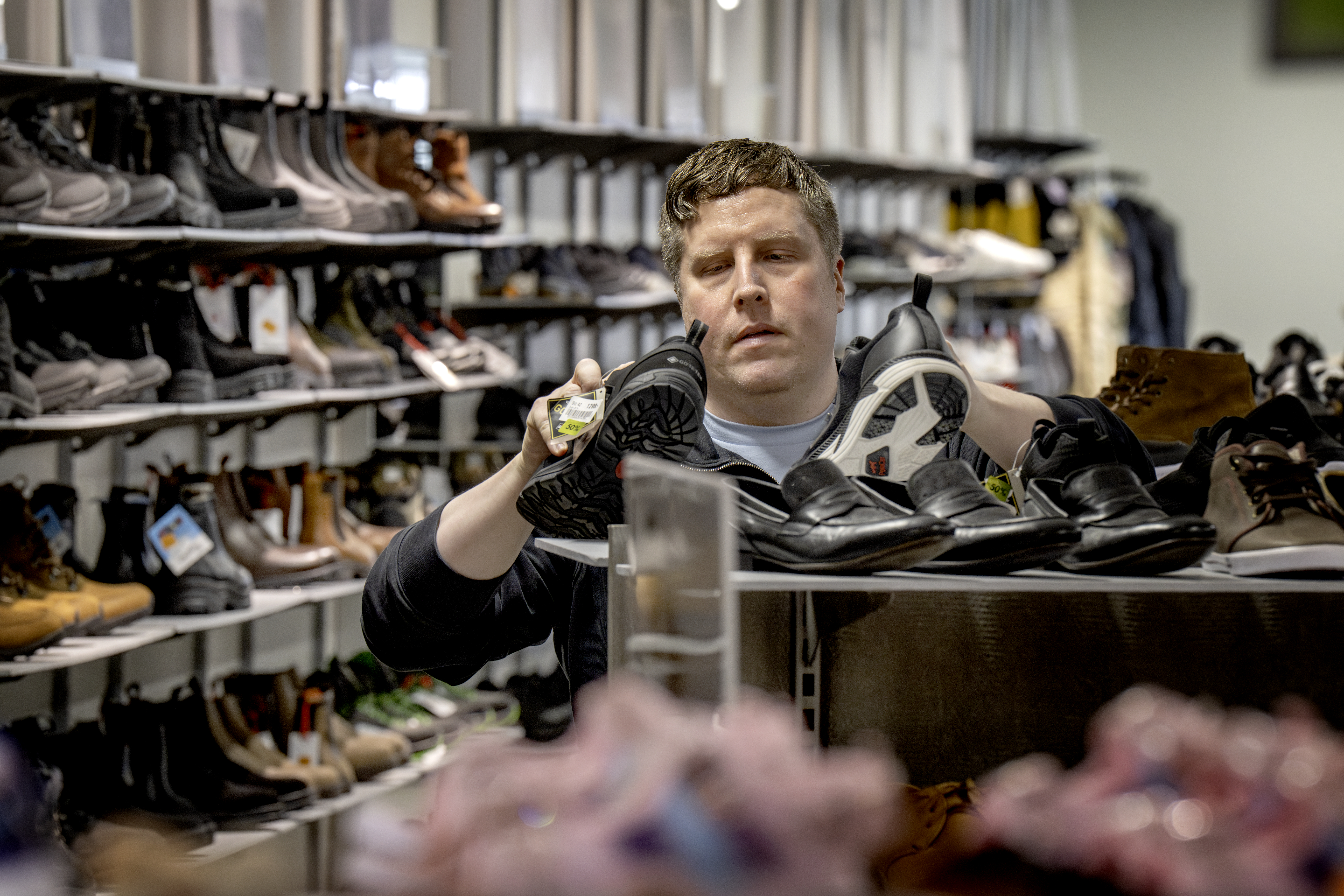 En person undersöker sulorna på två par skor i en butik, omgiven av andra skor på hyllor och ett bord.