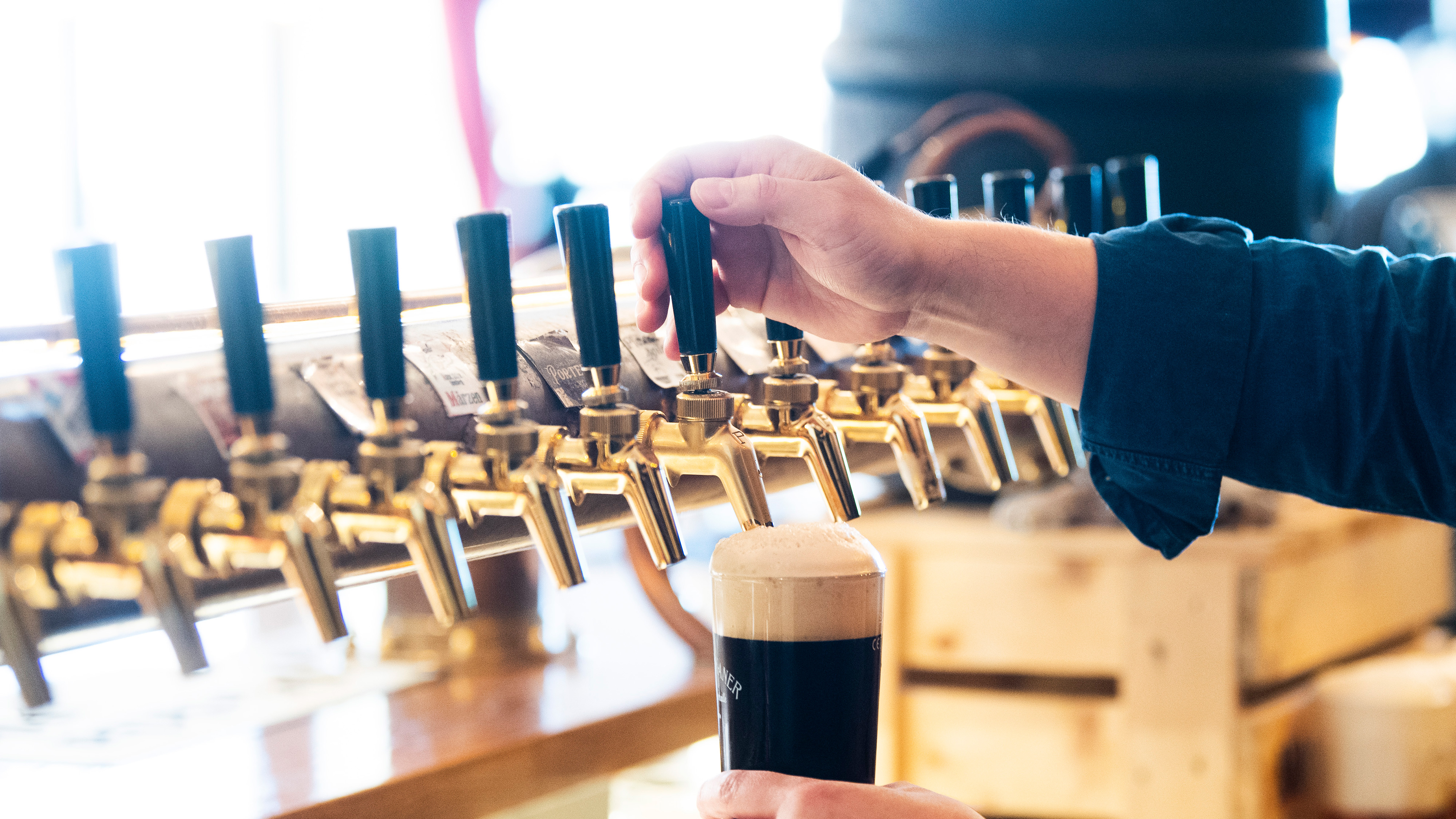 En person häller upp mörk öl i glas från en rad ölkranar på en bar.
