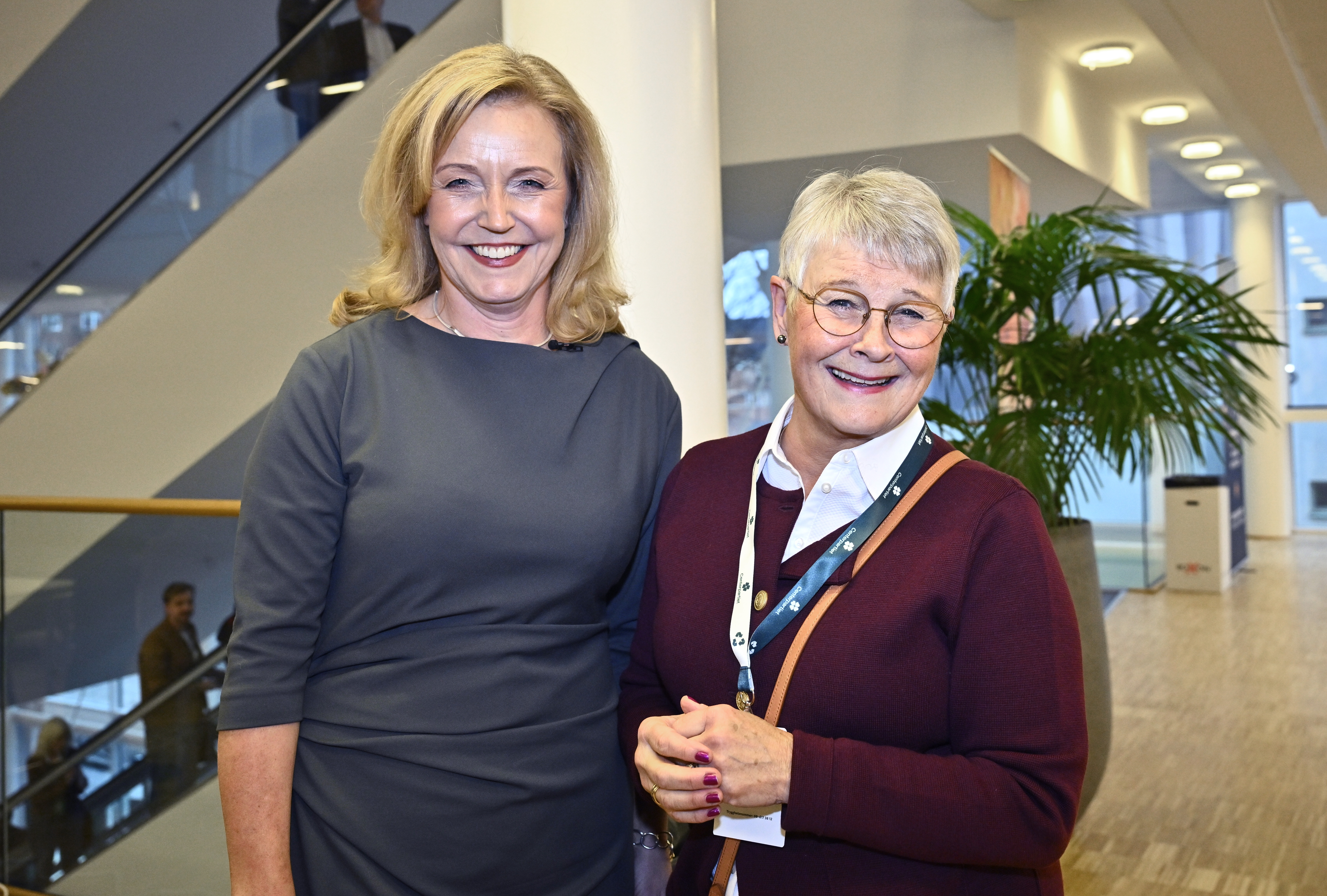 Två äldre kvinnor står inomhus och ler mot kameran i en byggnad med trappa, växt och personer i bakgrunden.