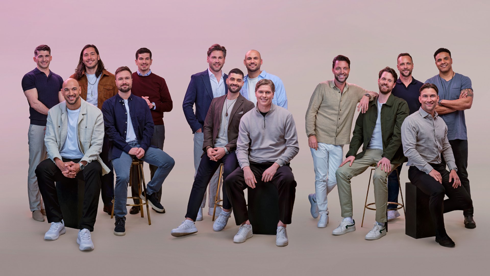 Fjorton män poserar tillsammans i halvformella kläder mot en neutral bakgrund, några står och några sitter på pallar eller block.