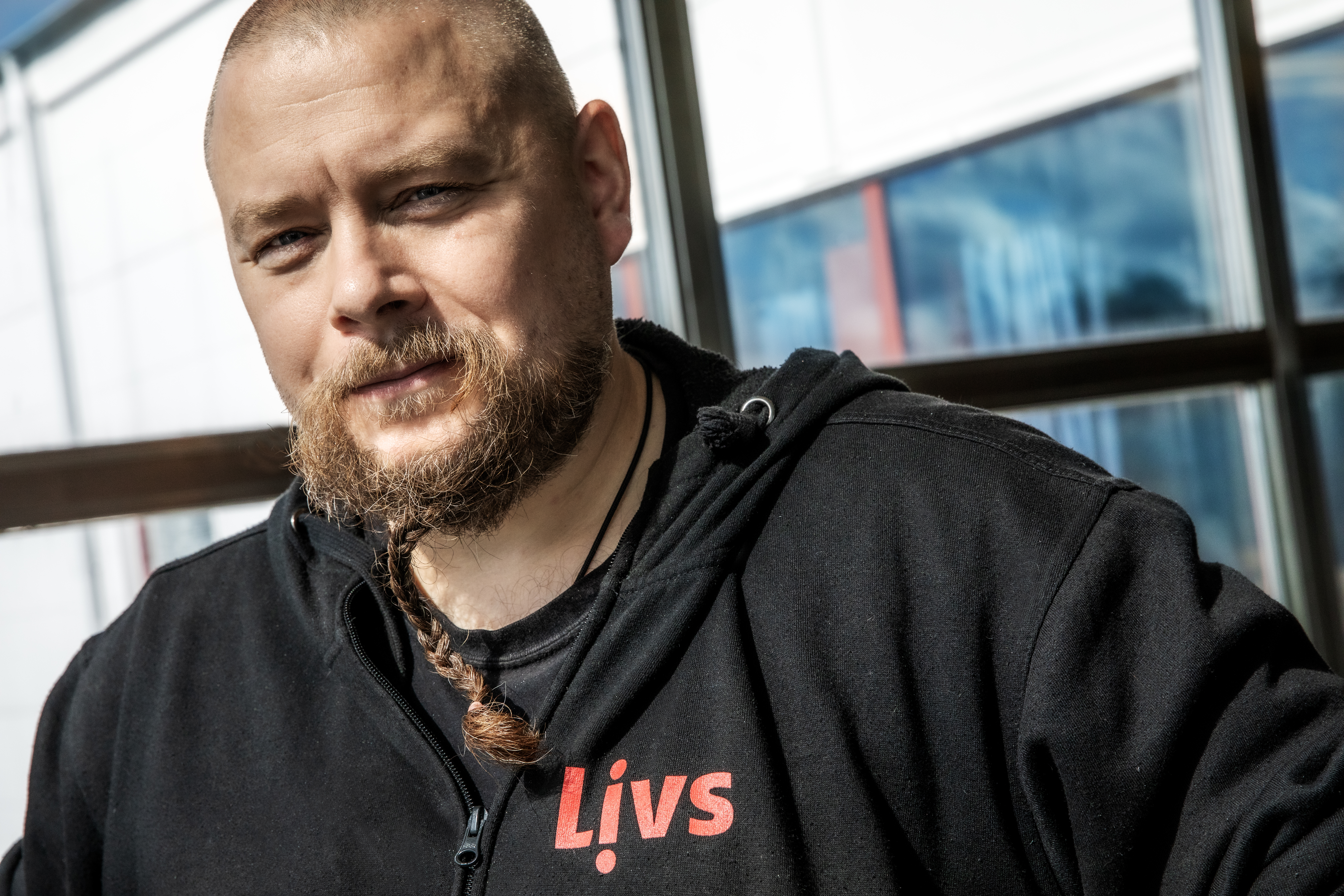En man med flätat skägg och rakat huvud har på sig en svart hoodie med texten "Livs" i rött, står inomhus vid ett fönster.