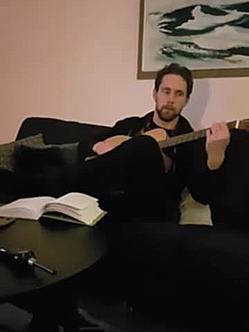 En man sitter på en soffa och spelar akustisk gitarr. Framför honom står ett bord med ett öppet anteckningsblock och en penna.