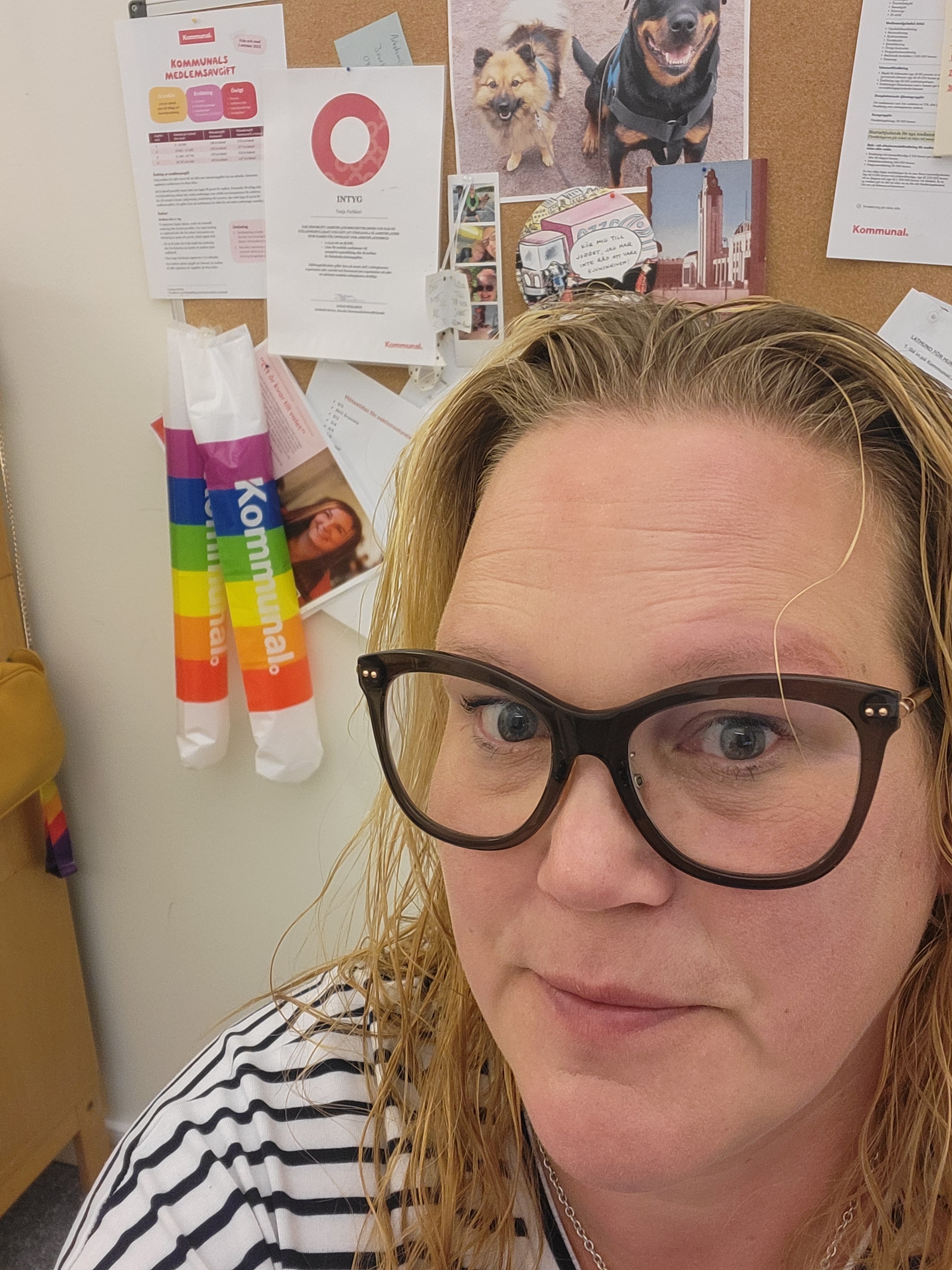 En kvinna med glasögon och blont, vått hår tar en selfie framför en anslagstavla med foton, lappar och en Kommunal-pryl i regnbågsfärger.