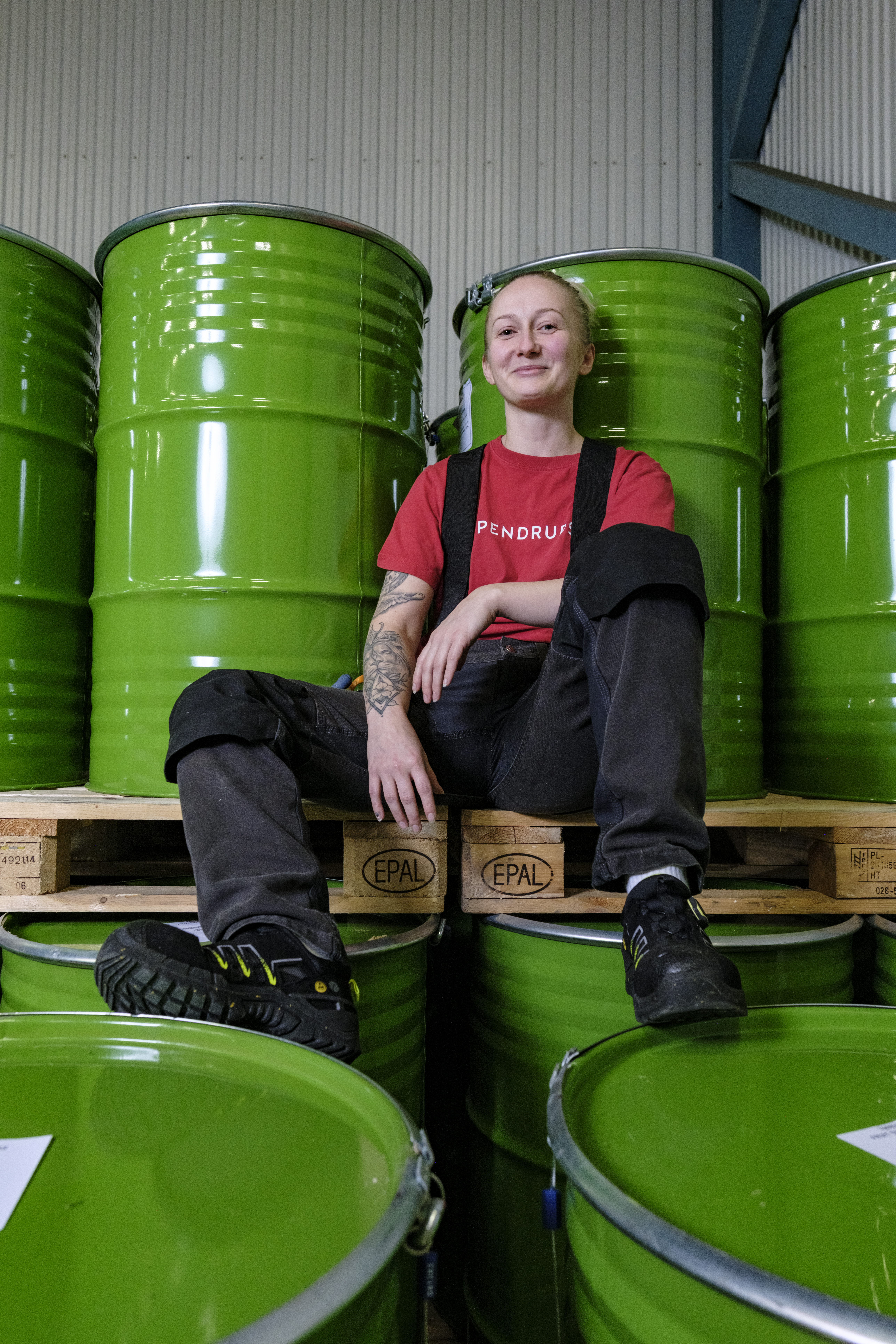 En person i arbetskläder sitter på en träpall mellan gröna industritunnor i ett lager och tittar mot kameran.