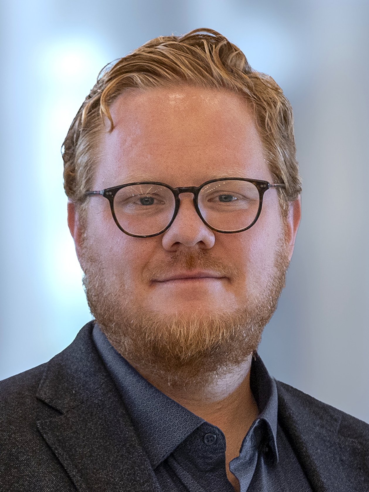 Emil Johansson med kort rött hår, skägg och glasögon bär en mörk kavaj och blå skjorta, står framför en suddig bakgrund.