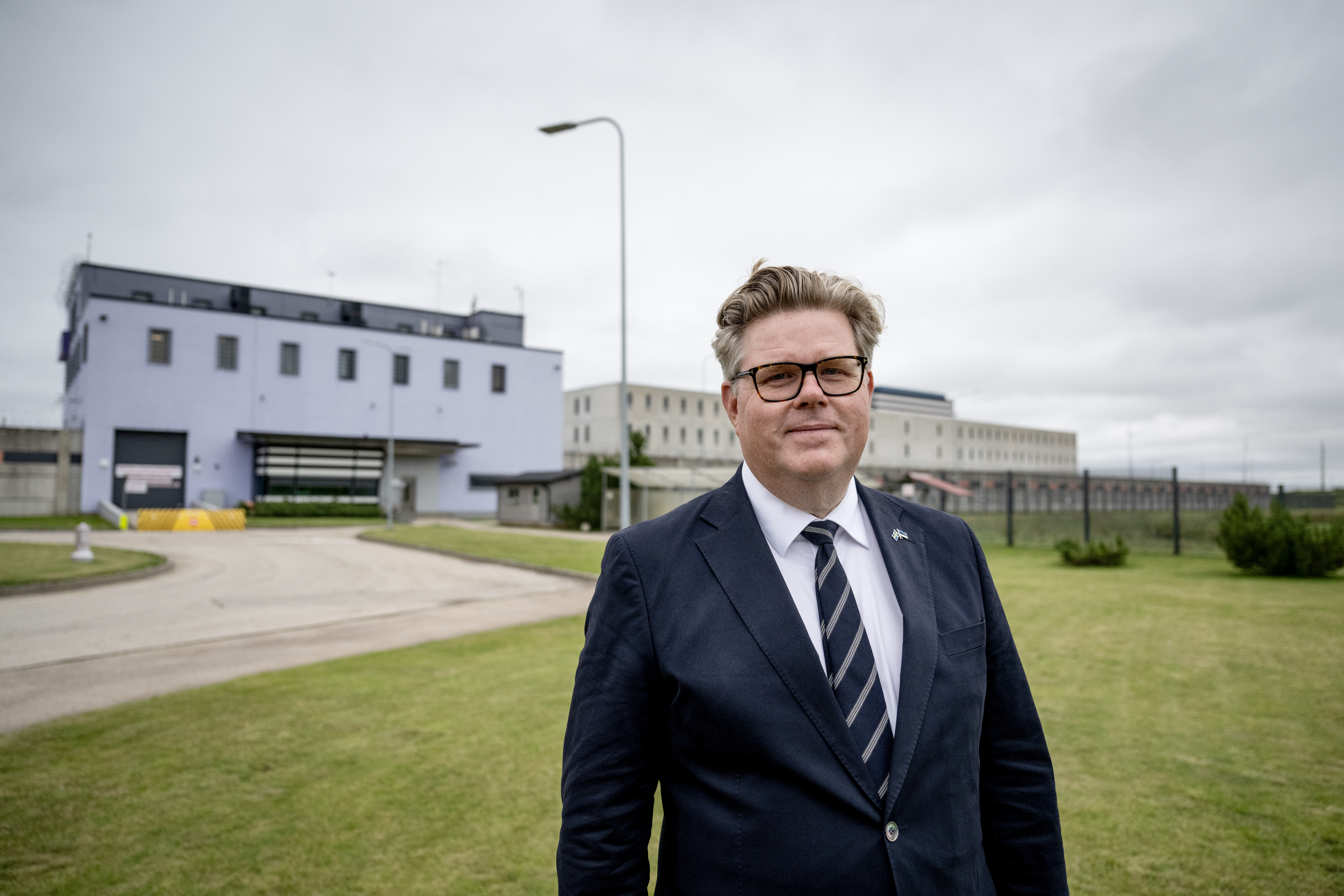 En man med kostym och glasögon står på gräs framför en stor byggnad med moln på himlen.