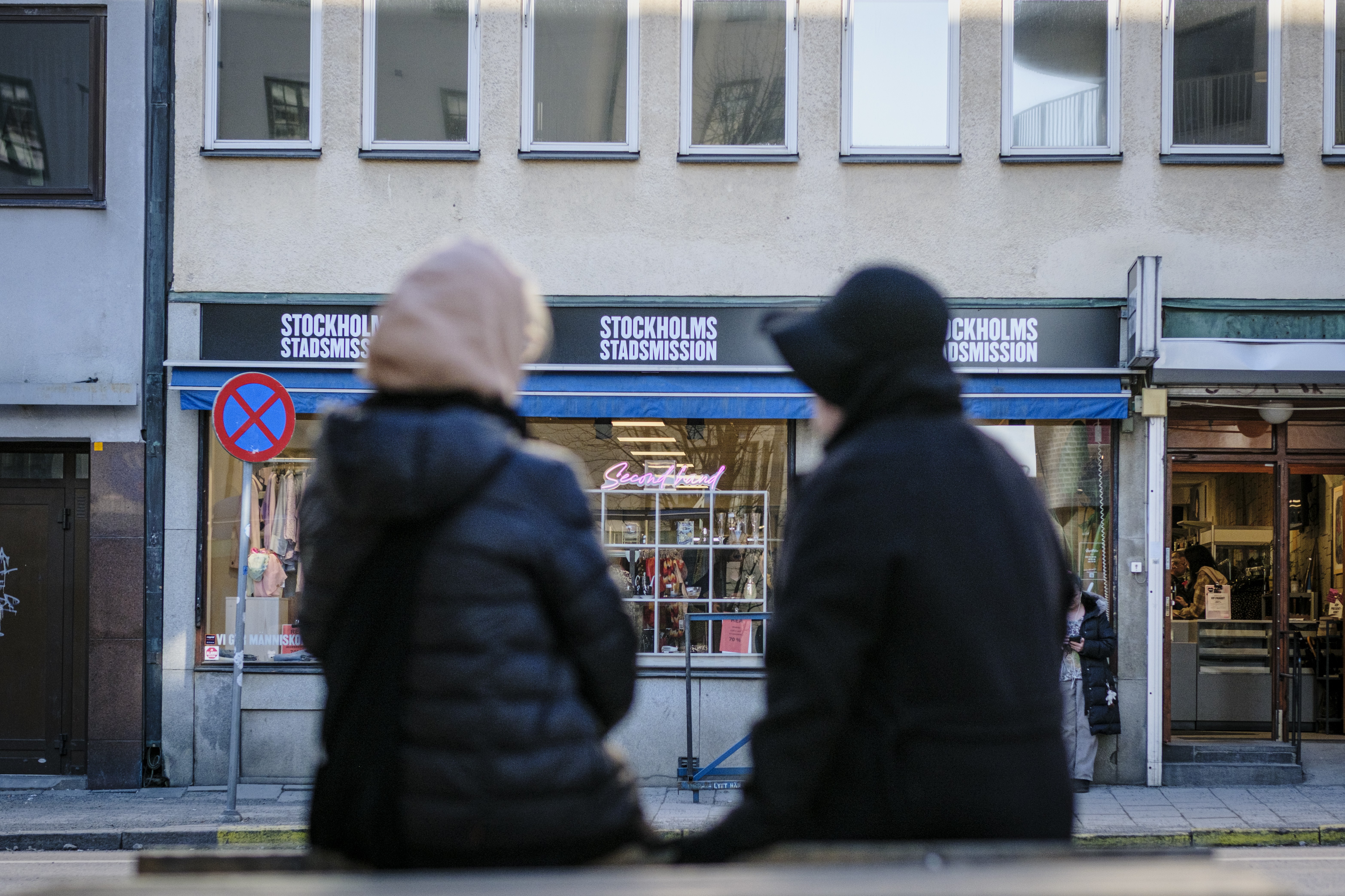 Två personer i vinterkläder sitter på en bänk vända mot en byggnad med skyltar där det står ”Stockholms Stadsmission” ovanför butiksfönstren.
