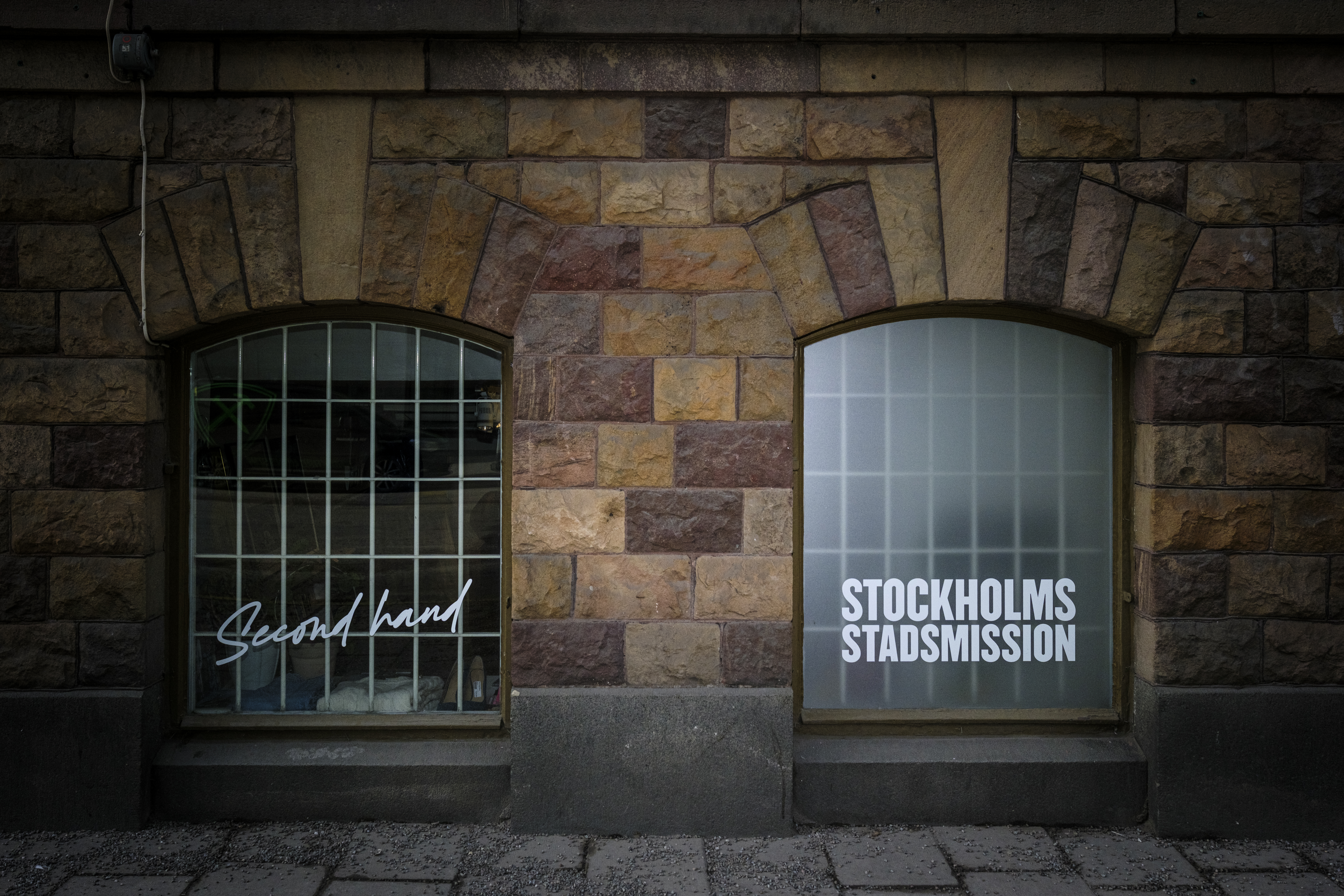 Två välvda fönster i en stenmur; på vänstra fönstret står "Second hand", på högra står "Stockholms Stadsmission".