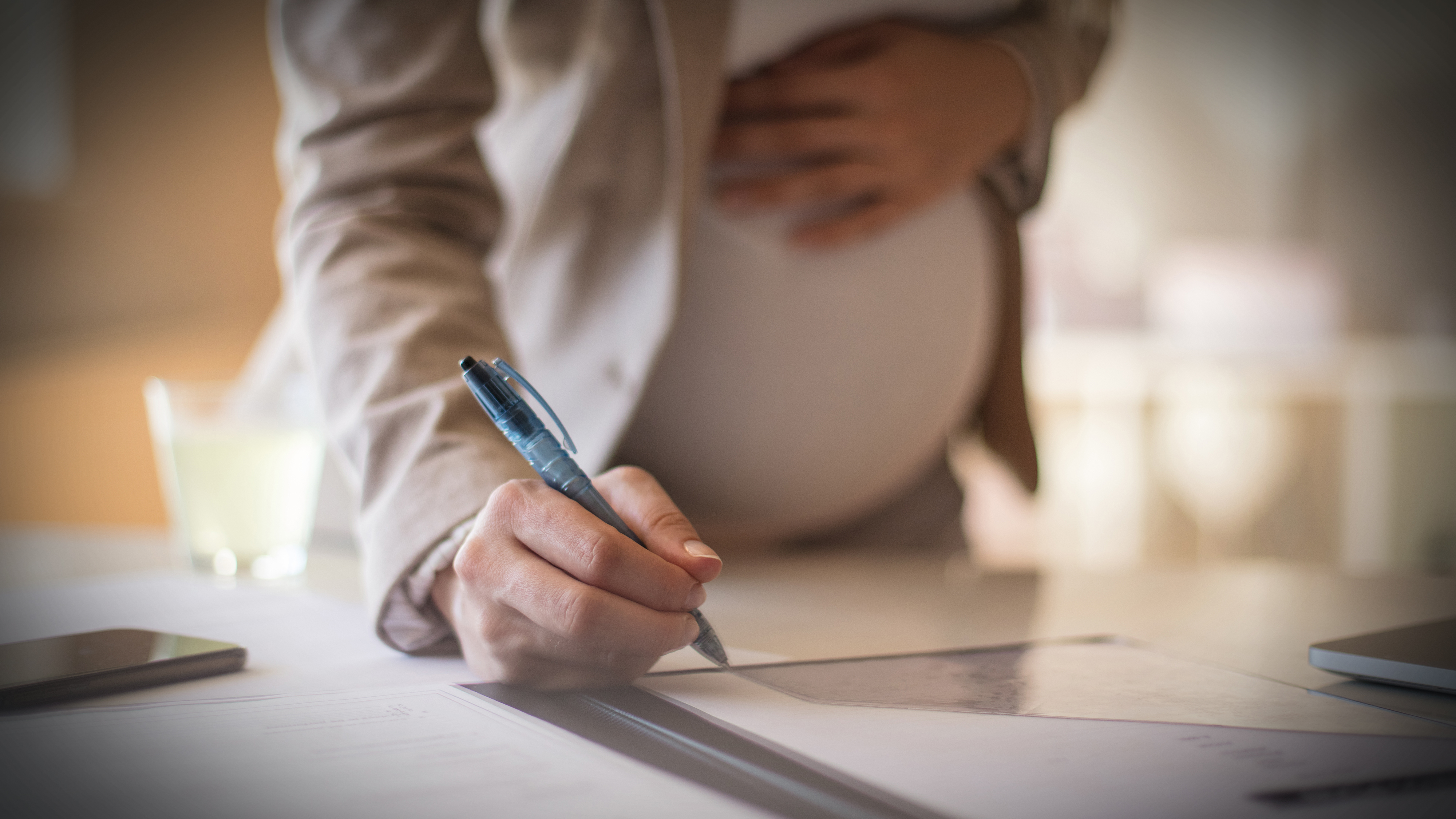 En gravid person i jobbkläder skriver under ett dokument vid ett skrivbord med ena handen på magen och den andra håller en penna.