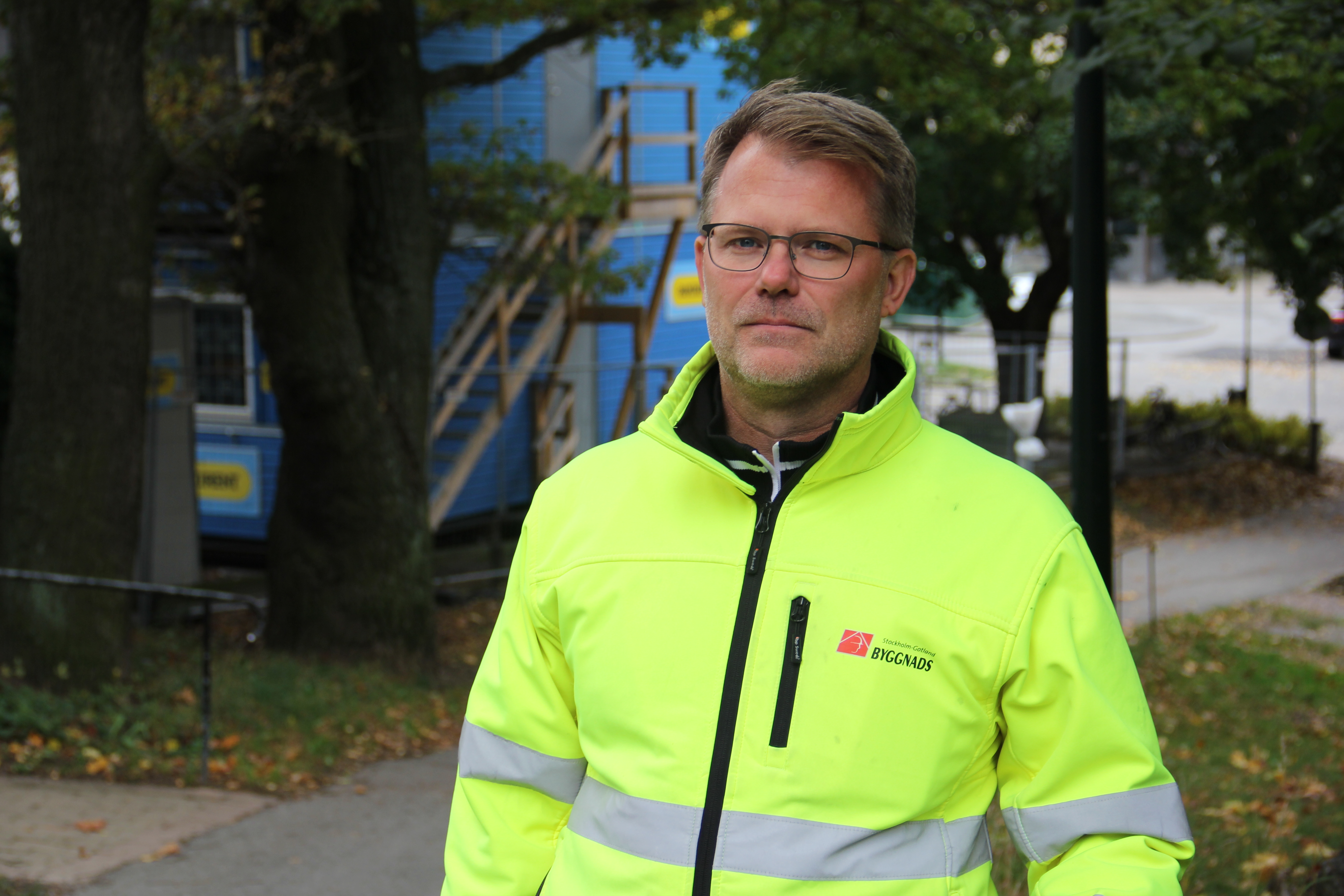 En man med glasögon och en gul varseljacka står utomhus nära träd och en byggarbetsplats.
