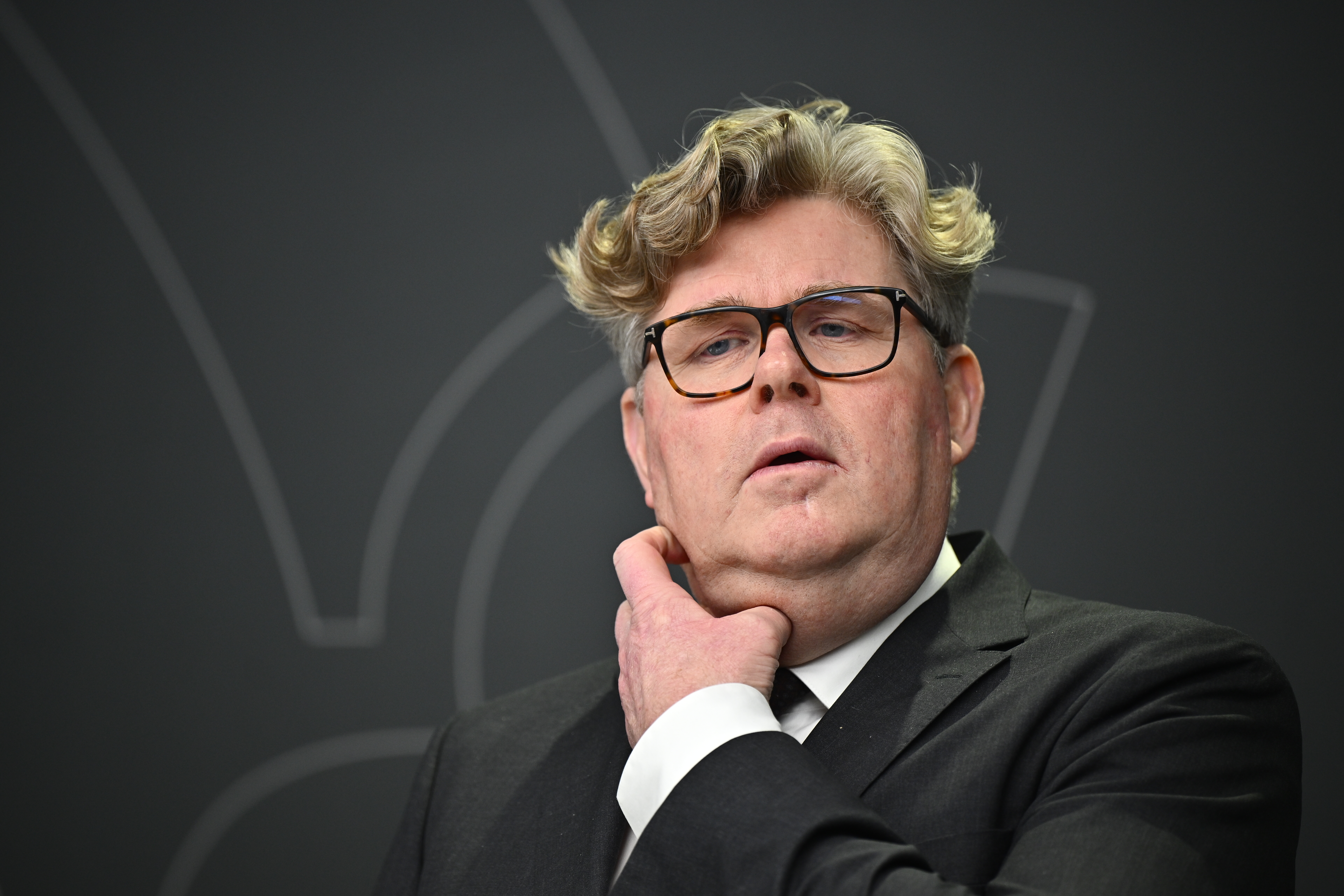 En man med kostym och glasögon står framför en mörk bakgrund, rör vid sin nacke och tittar lite uppåt.