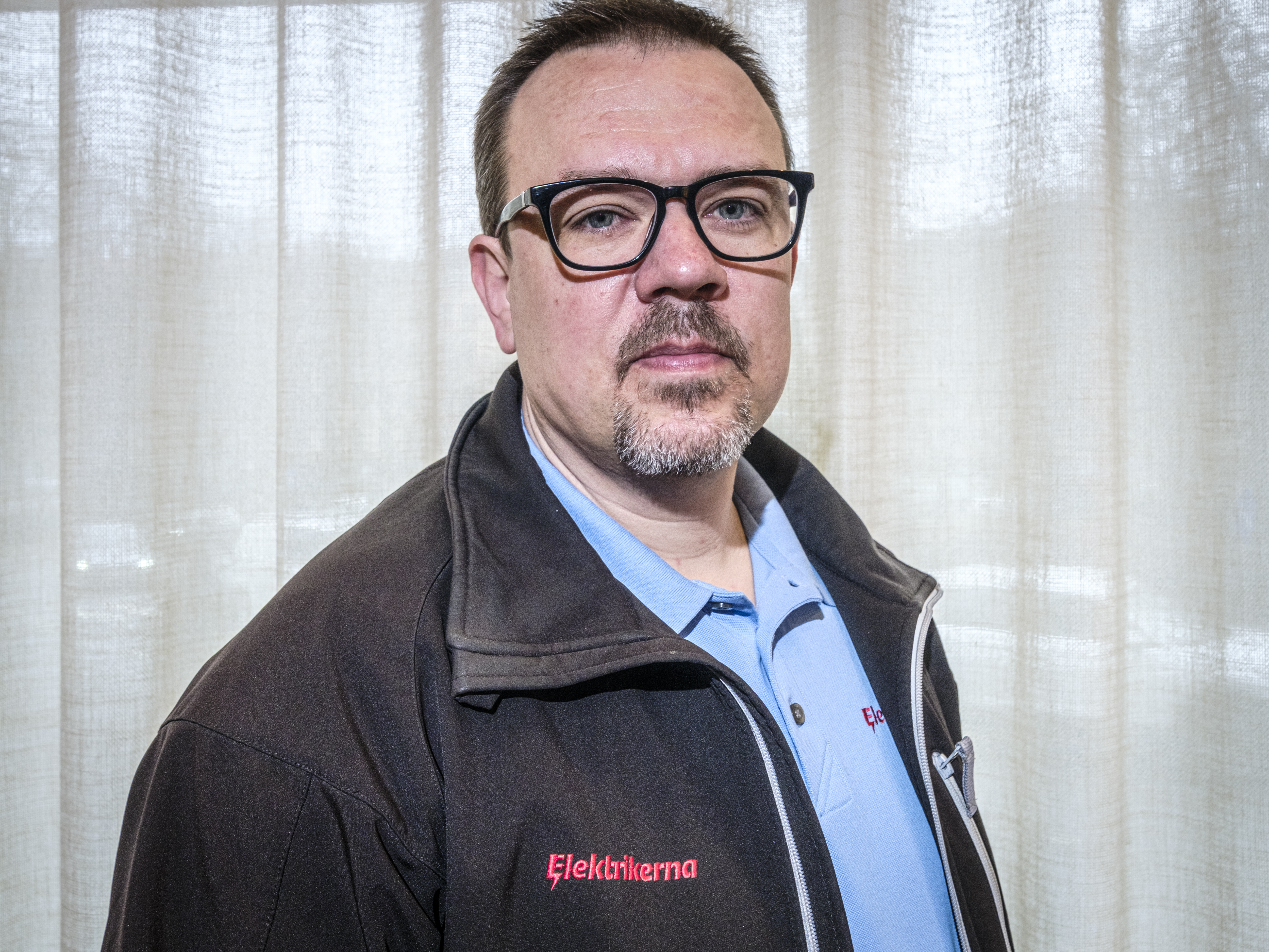 En man med kort hår, glasögon och getskägg står framför ett draperi, iförd ljusblå skjorta och mörk jacka med "Elektrikerna".