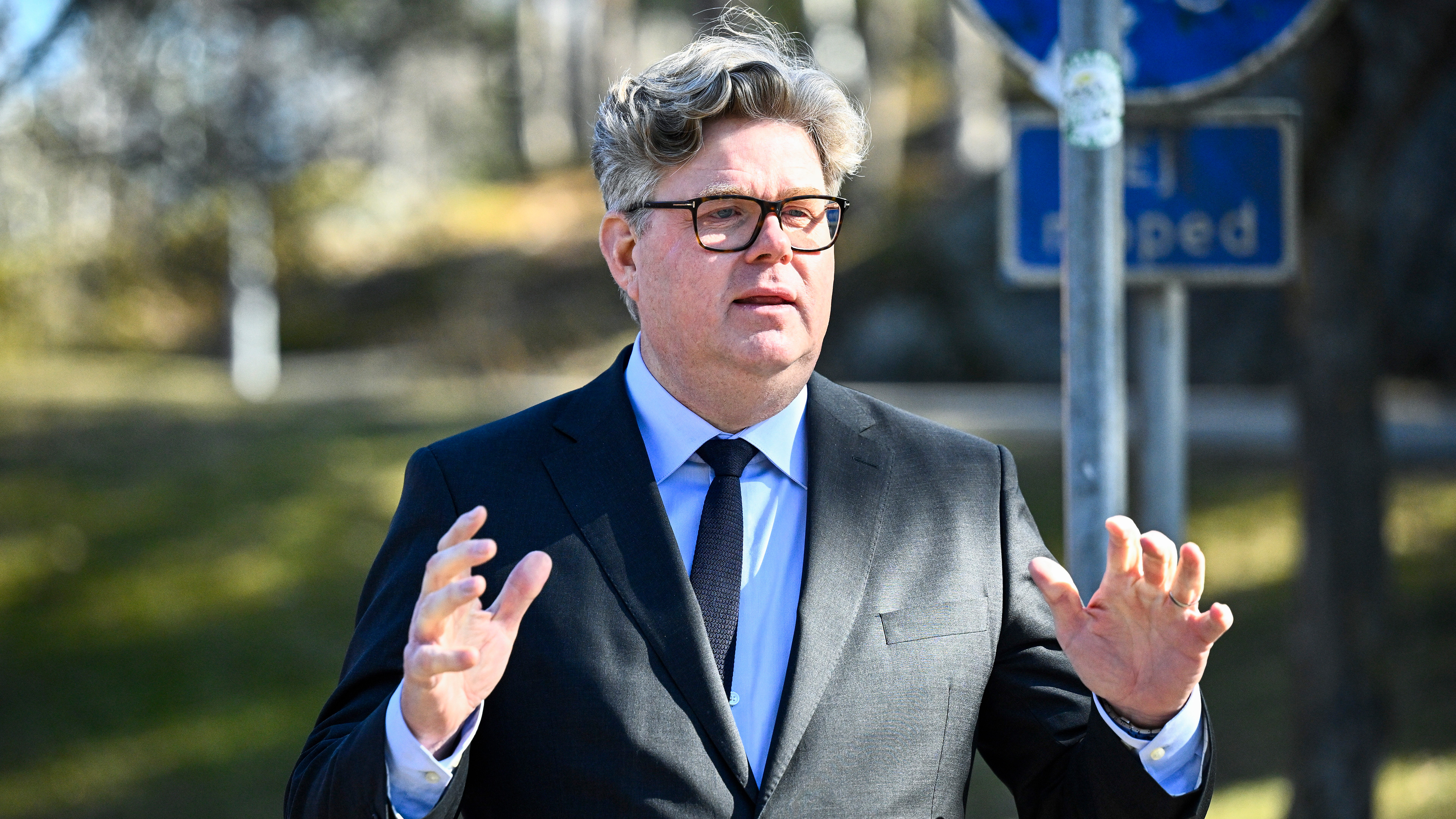 En man med kostym, slipsar och glasögon pratar utomhus och gestikulerar med båda händerna. En blå trafiksymbol syns bakom.