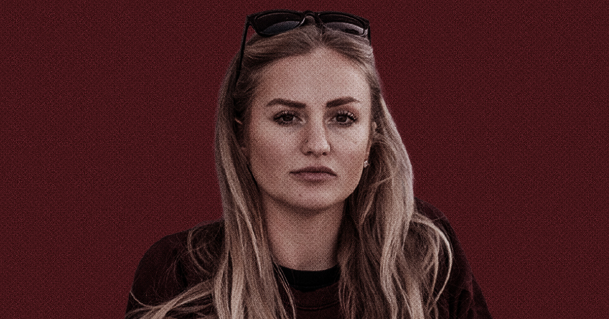 En kvinna med långt blont hår, mörk topp och solglasögon på huvudet sitter mot en enfärgad vinröd bakgrund.