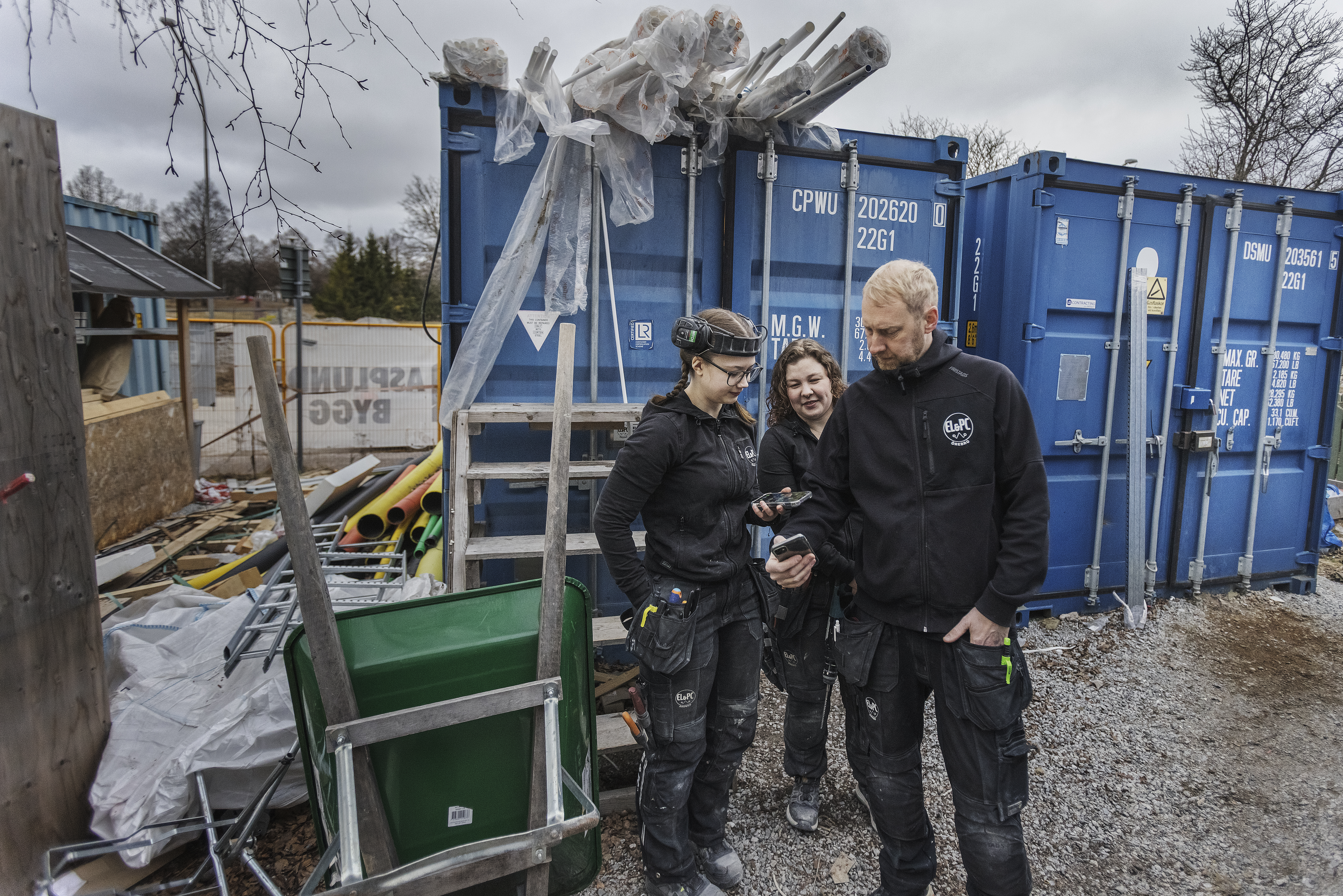 Två kvinnor och en man står på en byggarbetsplats fylld med fraktcontainrar och tittar på en mobiltelefon som hålls av en arbetare.