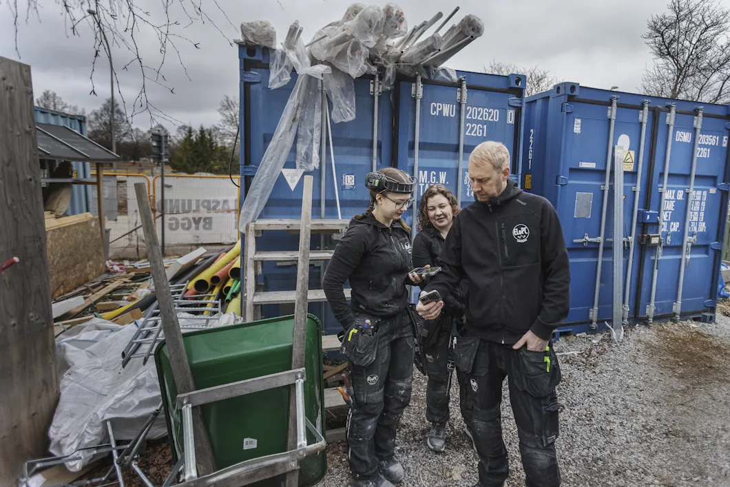 Två kvinnor och en man står på en byggarbetsplats fylld med fraktcontainrar och tittar på en mobiltelefon som hålls av en arbetare.