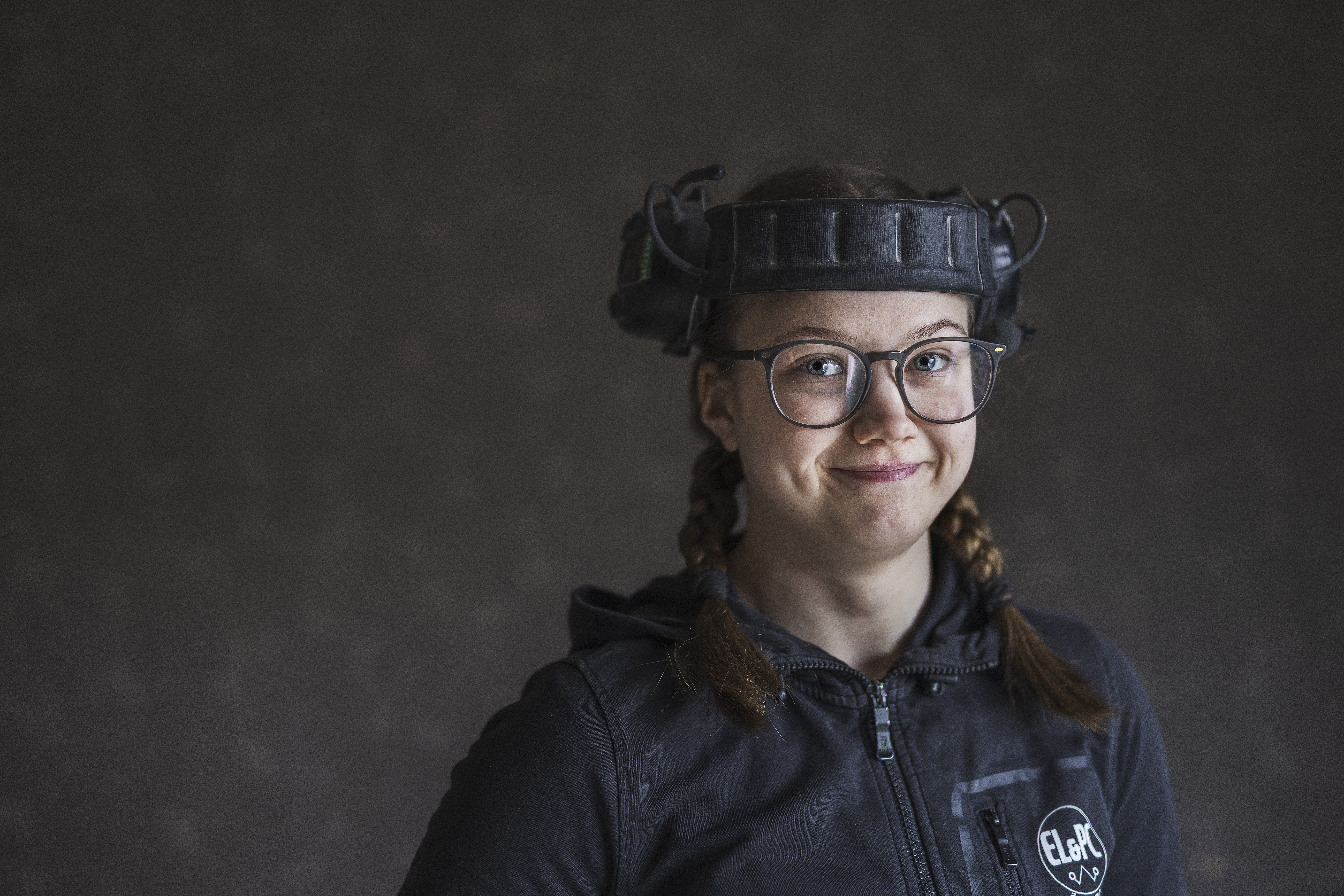 En person med glasögon och flätat hår och hörselkåpor på huvudet står mot en enkel, suddig bakgrund.