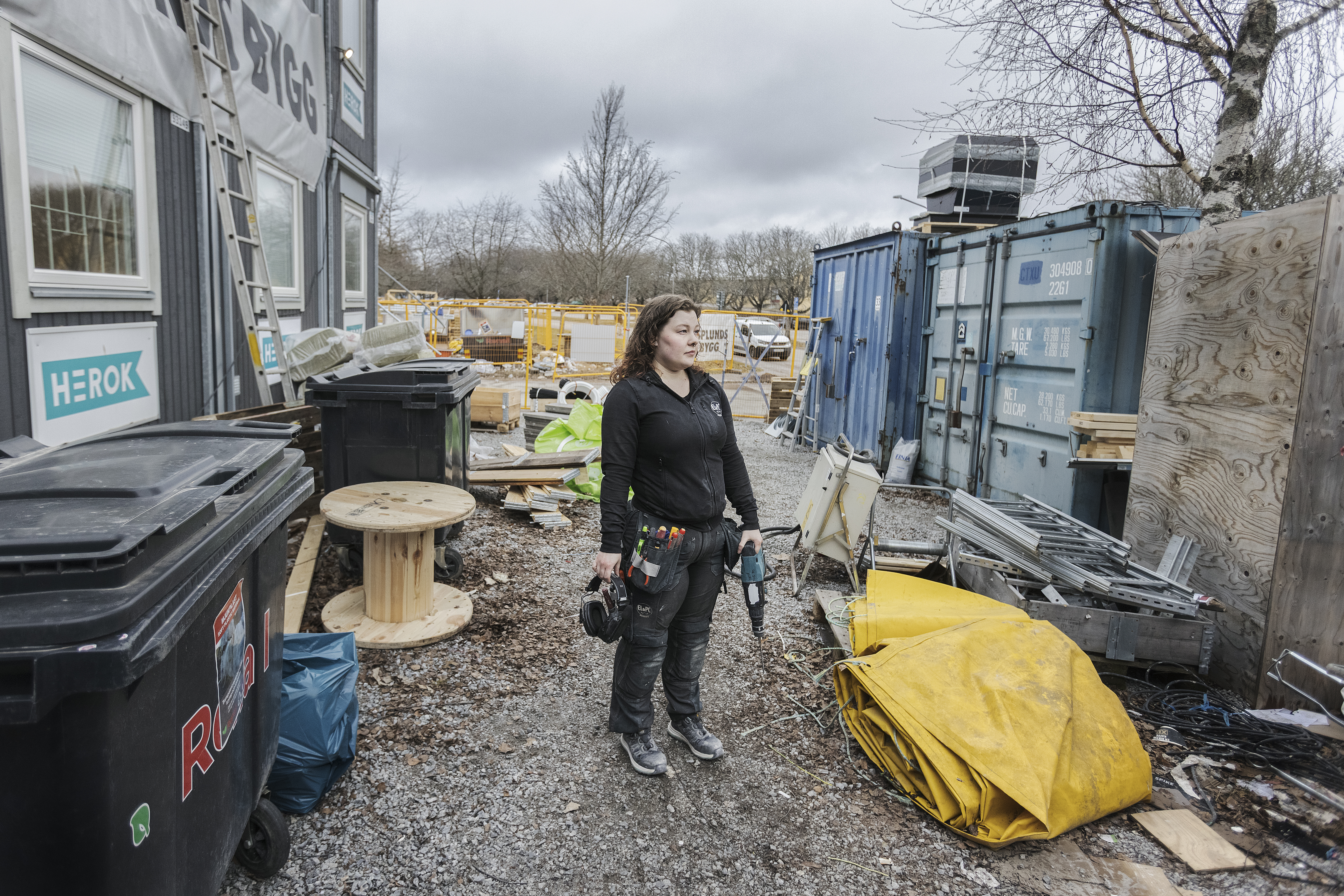 En person håller ett elverktyg i handen på en byggarbetsplats omgiven av utrustning, containrar, skräp och byggmaterial under en molnig himmel.