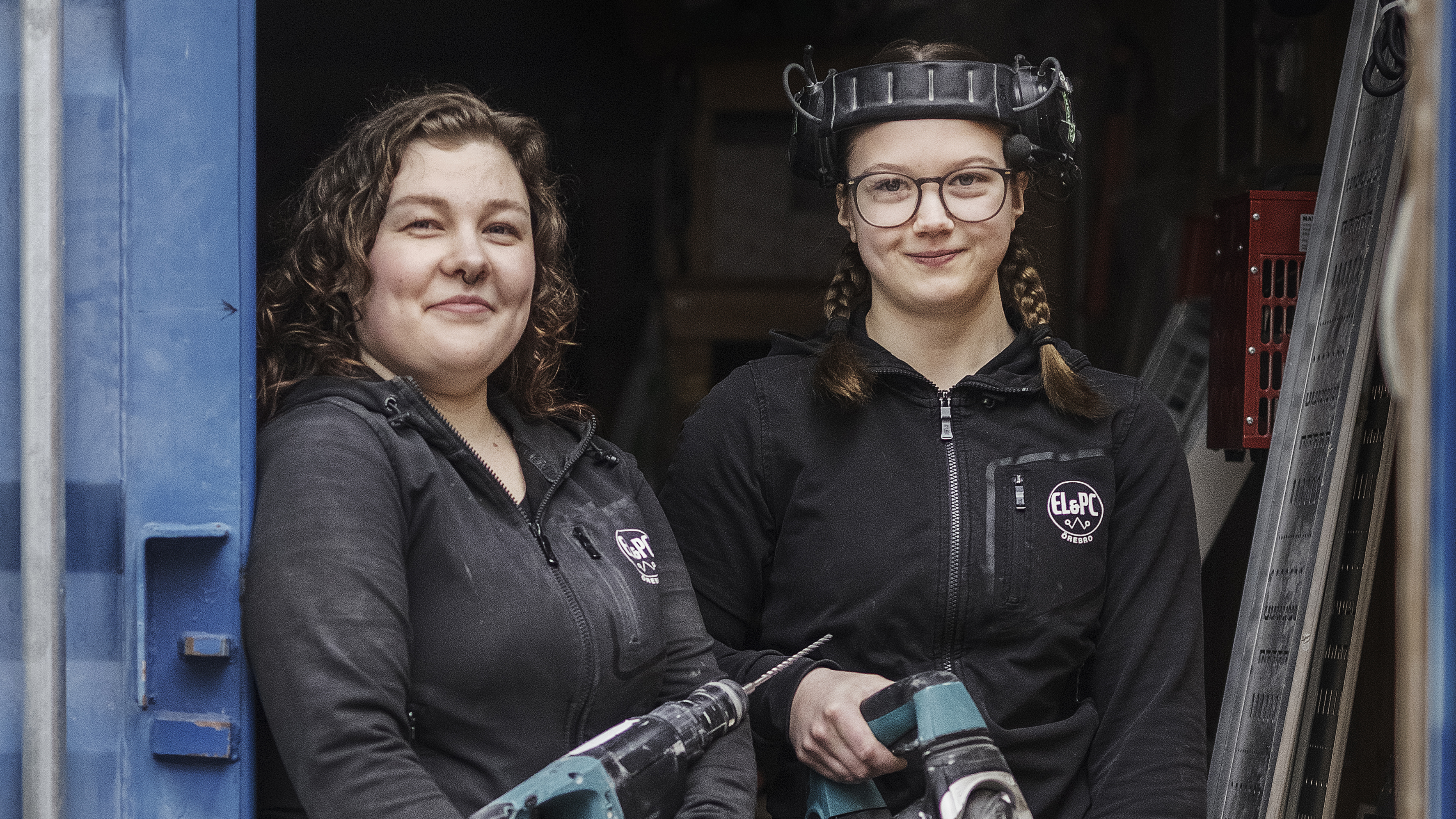 Två kvinnor i svarta arbetskläder står sida vid sida med elverktyg. En person bär en ansiktsskärm på huvudet. de är i verkstaden.