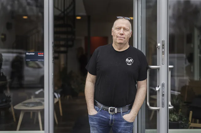 En man klädd i svart t-shirt och jeans står framför en glasdörr med händerna i fickorna. Inomhusmiljön syns bakom honom.