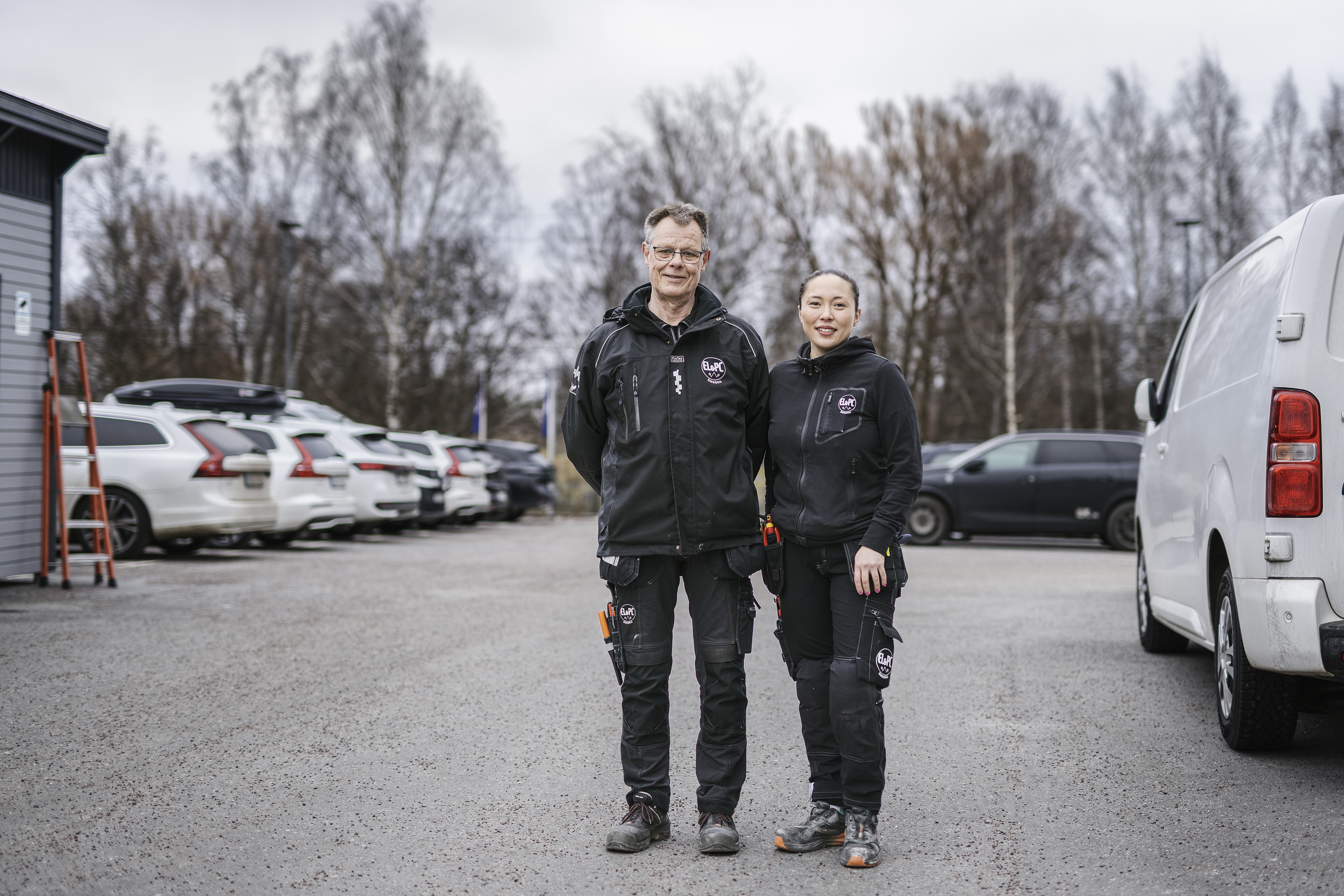 Två personer i svarta arbetskläder står sida vid sida på en parkeringsplats med flera bilar och bladlösa träd i bakgrunden.