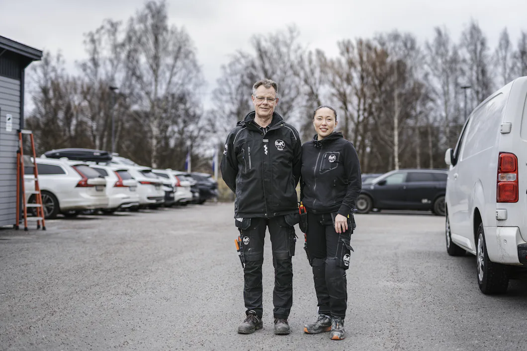Två personer i svarta arbetskläder står sida vid sida på en parkeringsplats med flera bilar och bladlösa träd i bakgrunden.
