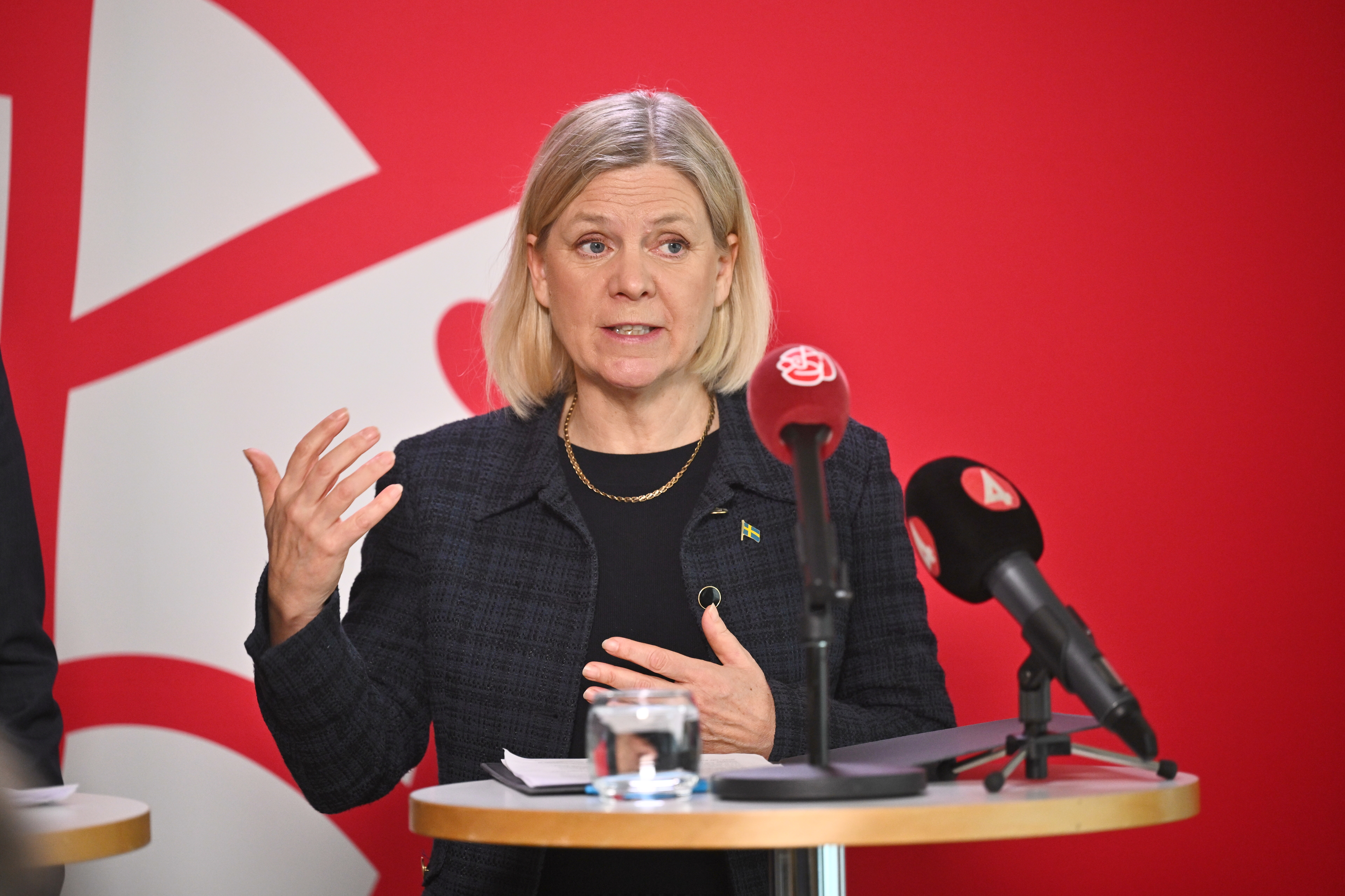 En kvinna står vid ett podium och talar i mikrofoner, gestikulerar med ena handen, framför en röd bakgrund med vit design.