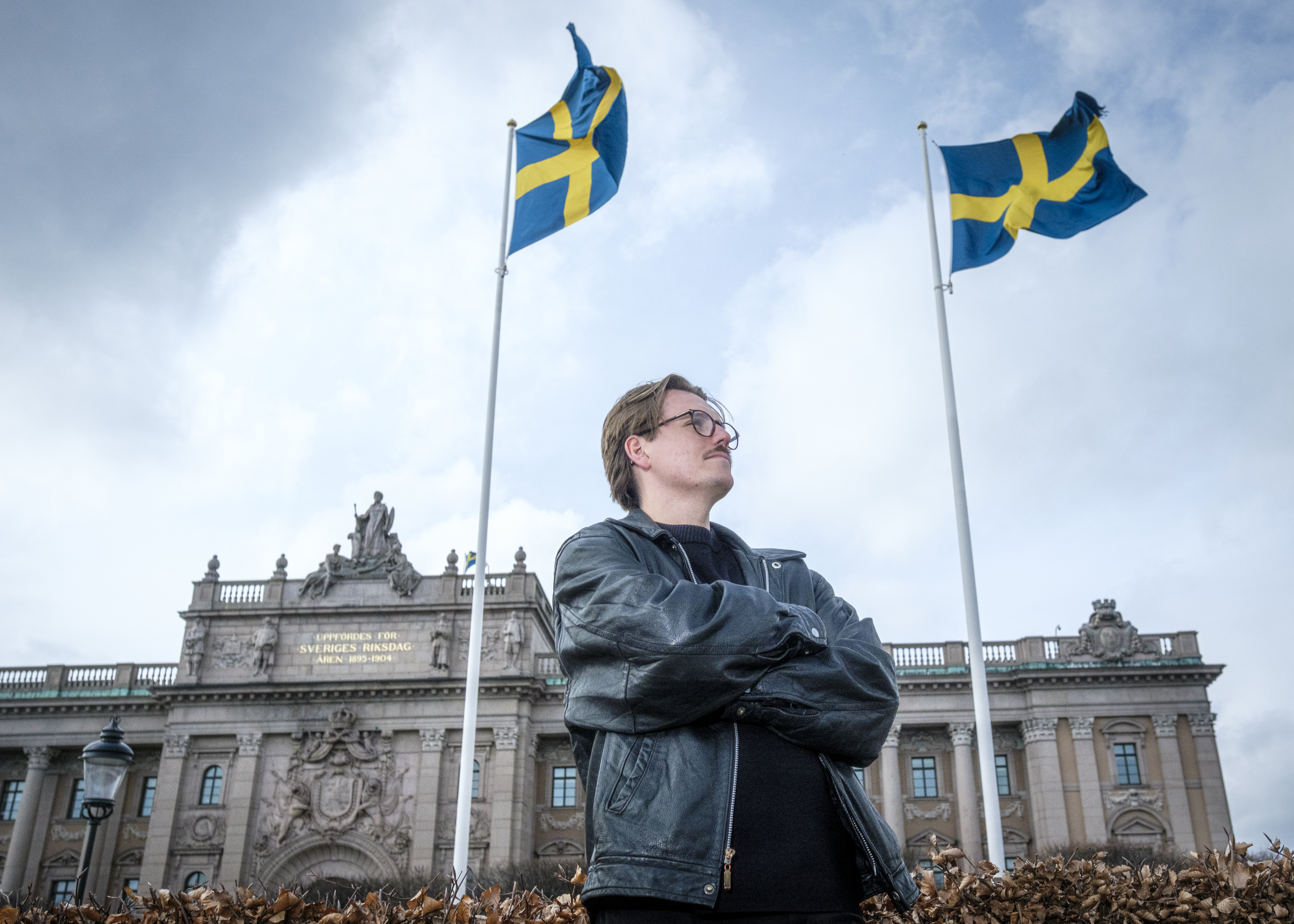 En person i svart skinnjacka står med korslagda armar framför en byggnad. Två svenska flaggor syns ovanför.