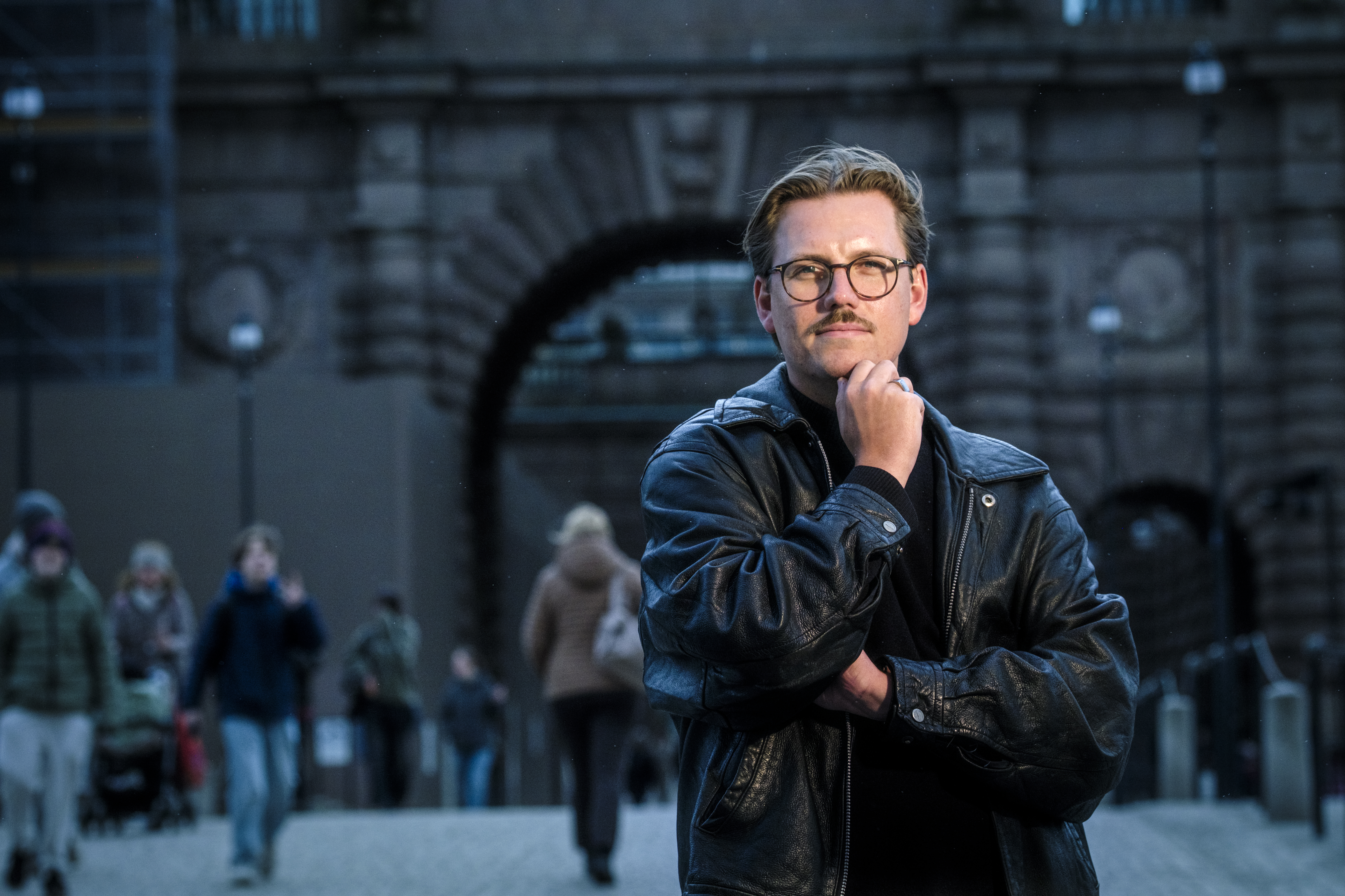 En man med svart skinnjacka och glasögon står med handen på hakan framför en stenbyggnad, medan folk går i bakgrunden.