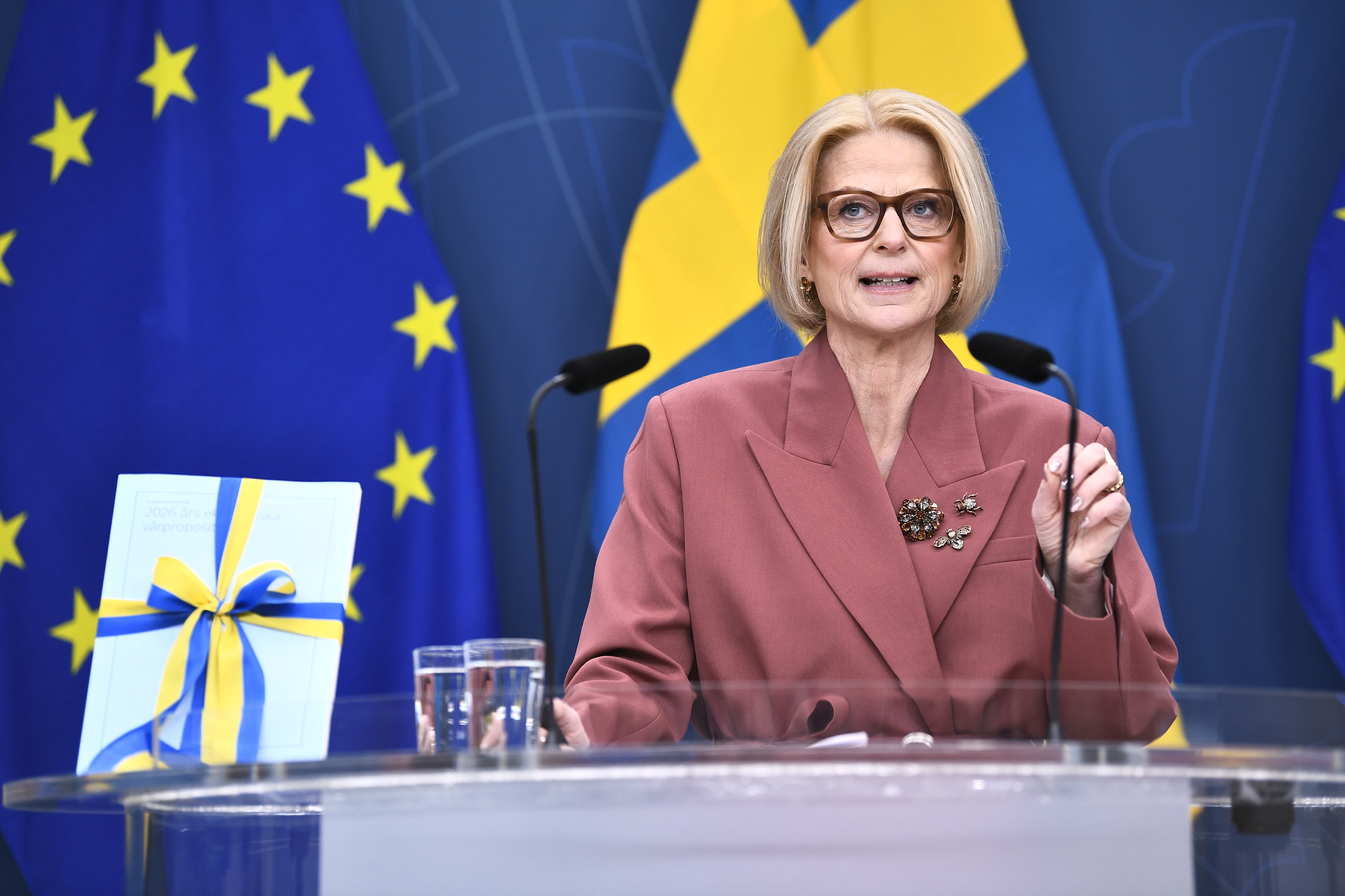 En kvinna i rosa dräkt talar vid en talarstol med EU:s och Sveriges flaggor bakom sig. Ett häfte med ett blågult band ligger på podiet.