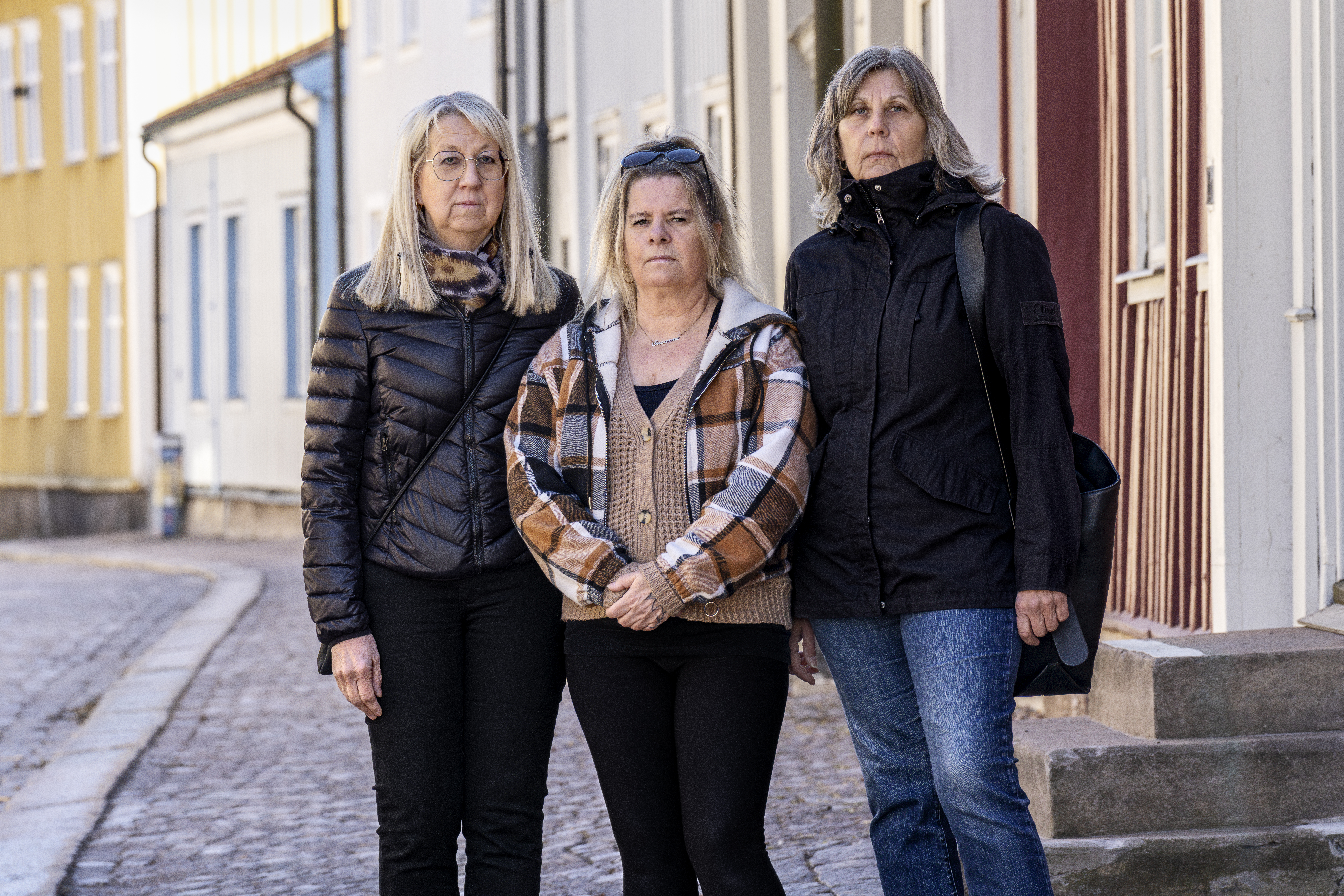 Tre medelålders kvinnor står bredvid varandra på en gatubeläggning, bär jackor och tittar mot kameran, byggnader syns bakom.