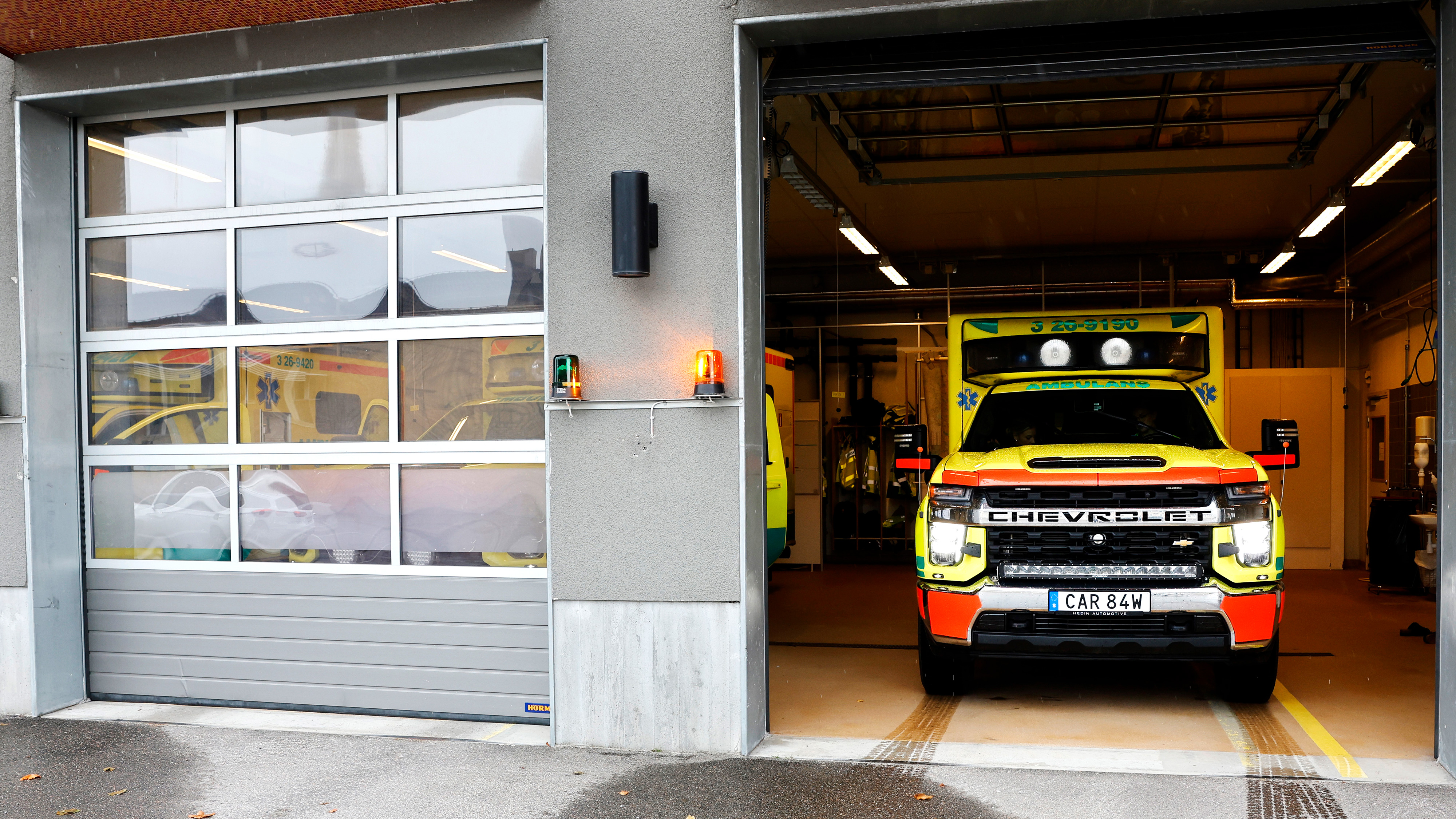 En gul och grön Chevrolet-ambulans står parkerad i ett garage med fronten utåt. En annan ambulans syns delvis till vänster bakom en glasdörr.