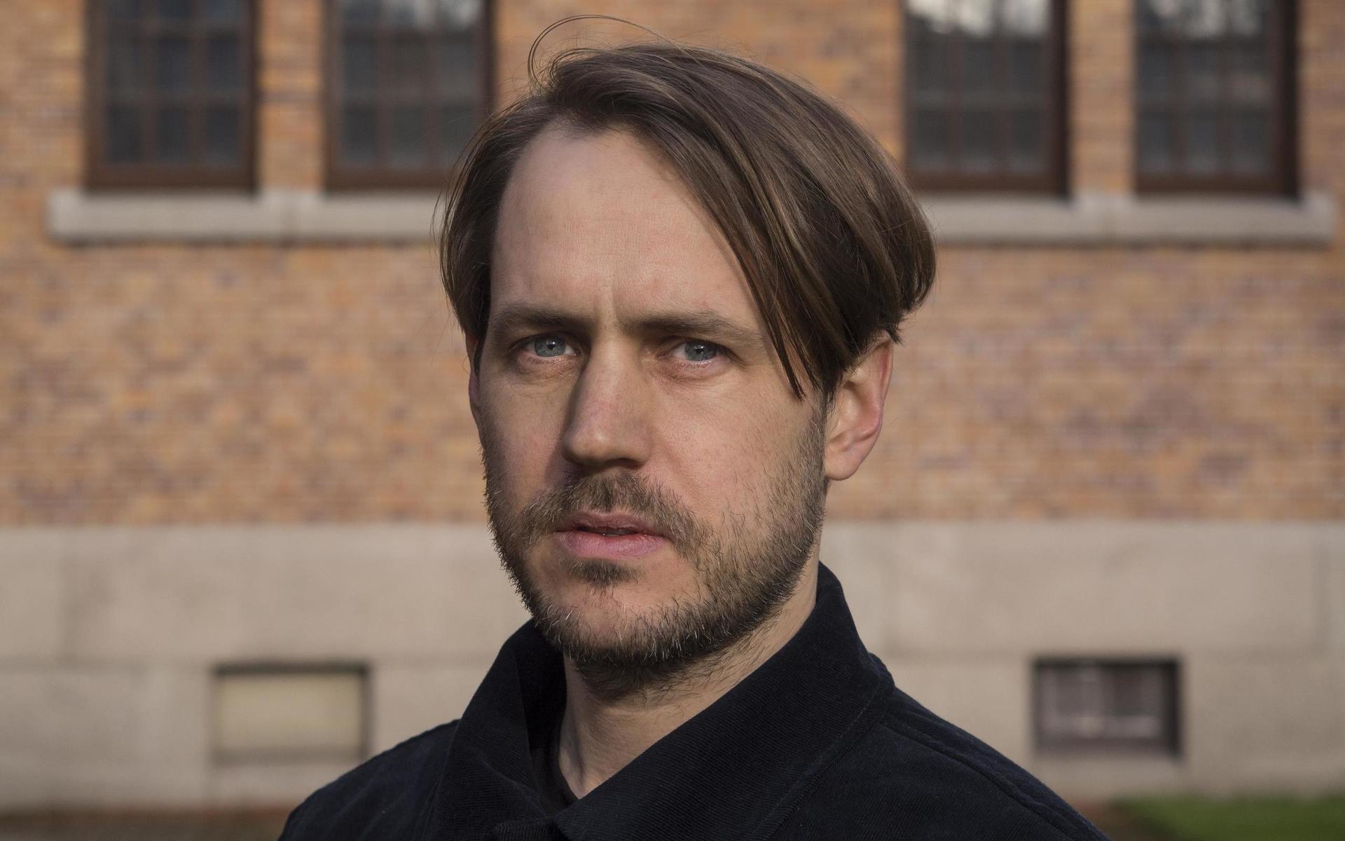 Johan Alfonsson, med ljusbrunt hår och kort skägg, står framför en tegelbyggnad och tittar in i kameran.