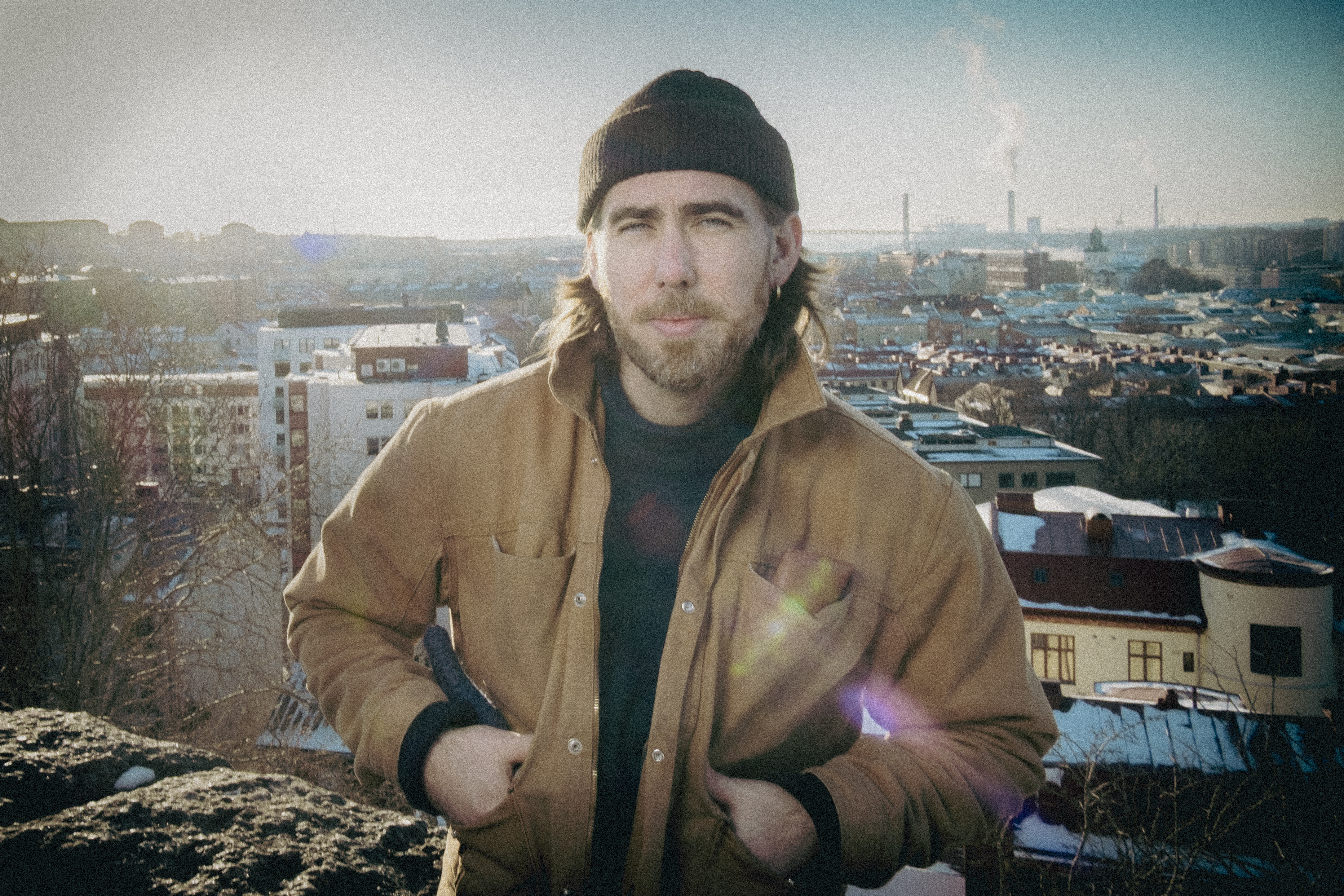 En person med brun jacka och svart mössa står utomhus med en stadssilhuett och fabriksrök i bakgrunden.