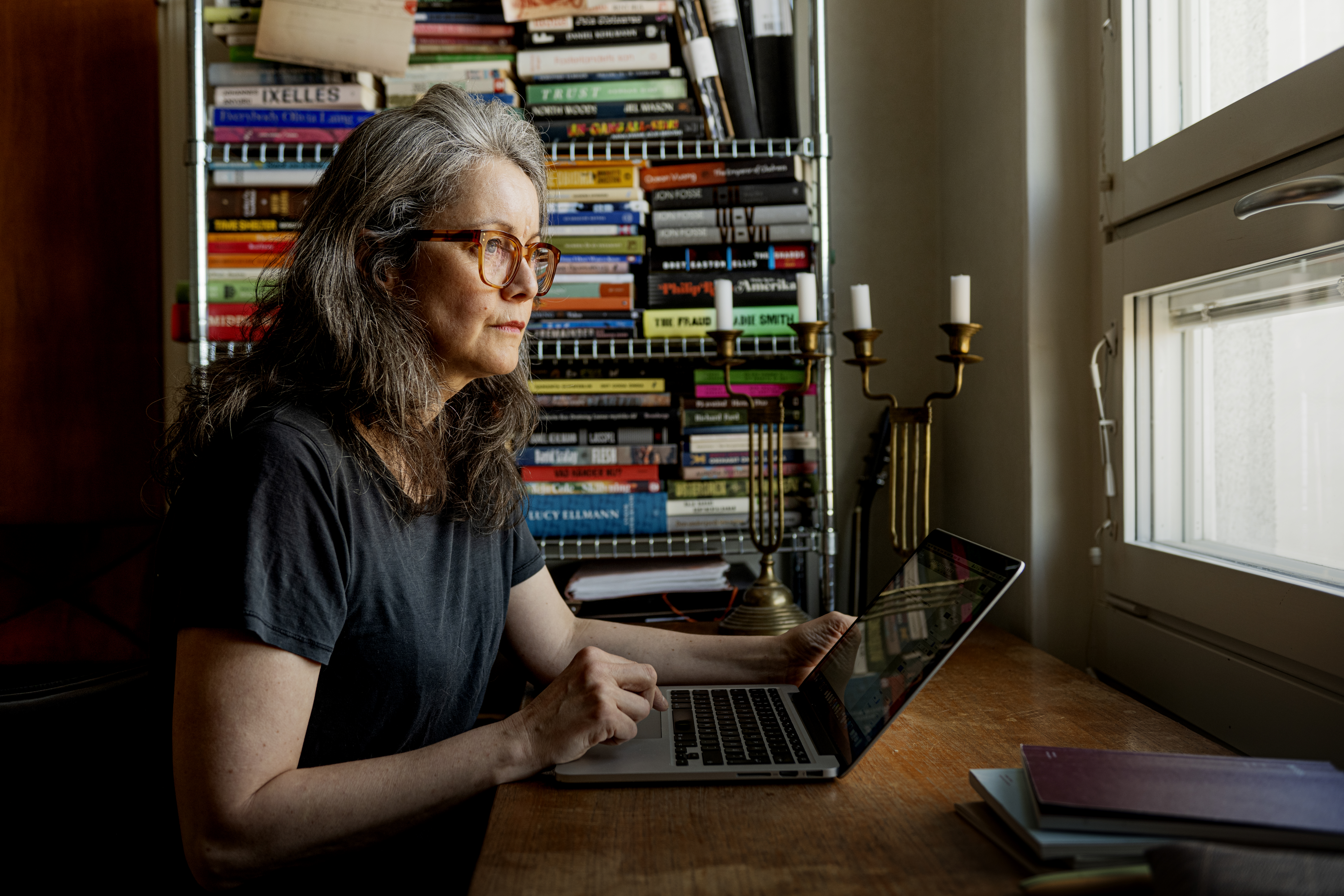 En kvinna med grått hår och glasögon sitter vid ett träbord och använder en bärbar dator. Bakom henne finns en bokhylla och ett fönster.