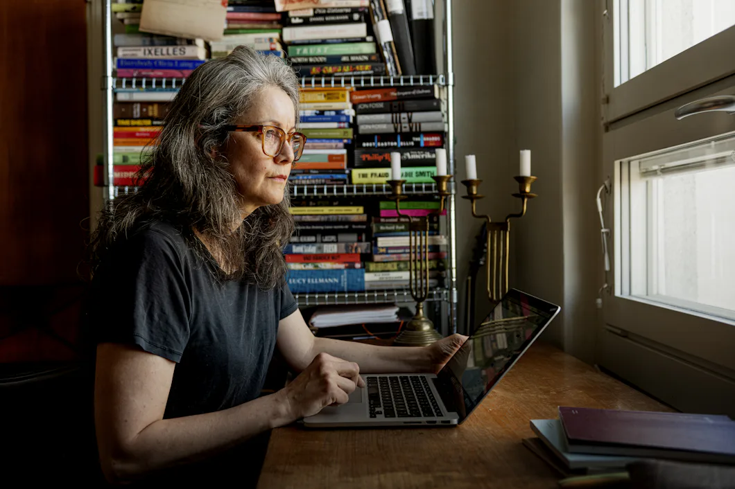En kvinna med grått hår och glasögon sitter vid ett träbord och använder en bärbar dator. Bakom henne finns en bokhylla och ett fönster.