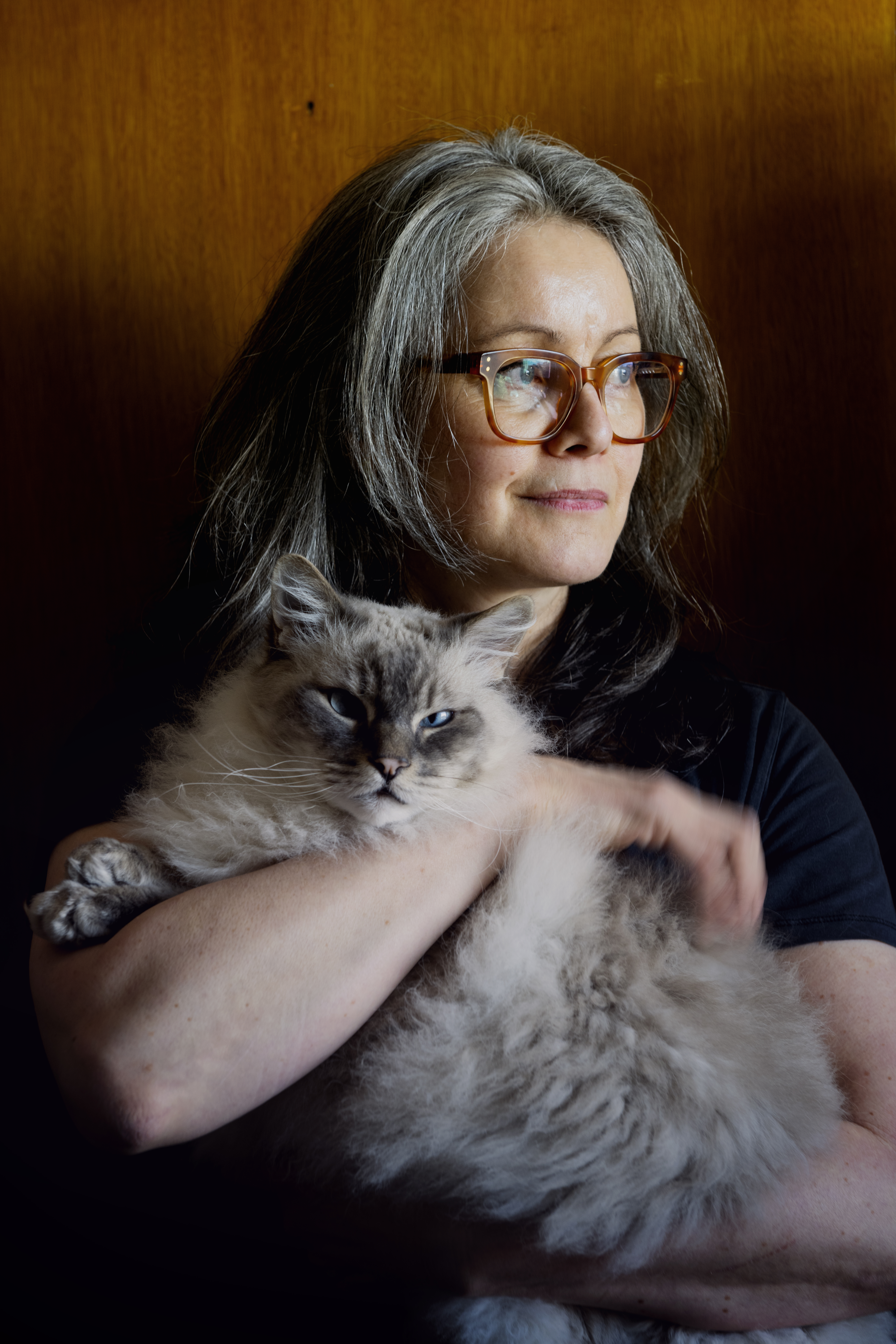 En kvinna med grått hår och glasögon håller i en grå katt och tittar lite åt sidan framför en bakgrund av trä.