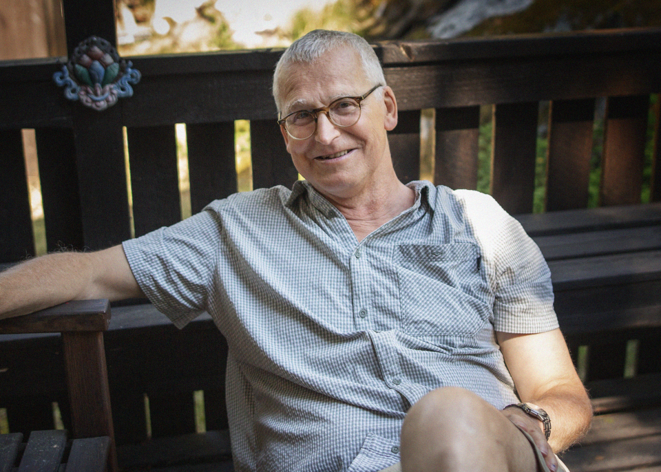 En äldre man med glasögon och kort grått hår sitter på en träbänk utomhus och ler mot kameran.
