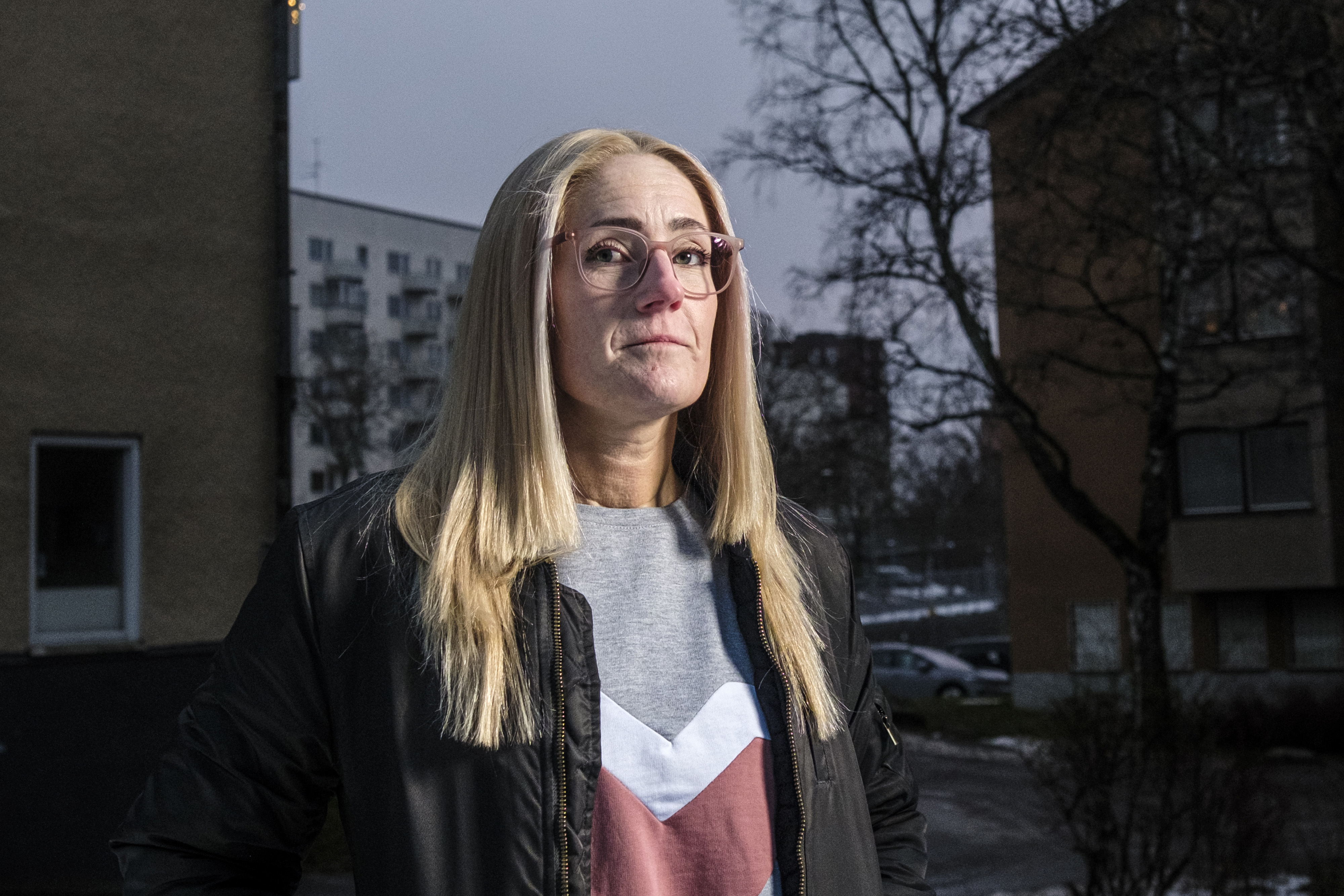 Karin Melin, med långt blont hår och glasögon, står utomhus i ett bostadsområde iklädd kavaj och tröja. Du kan se hus och träd i bakgrunden.